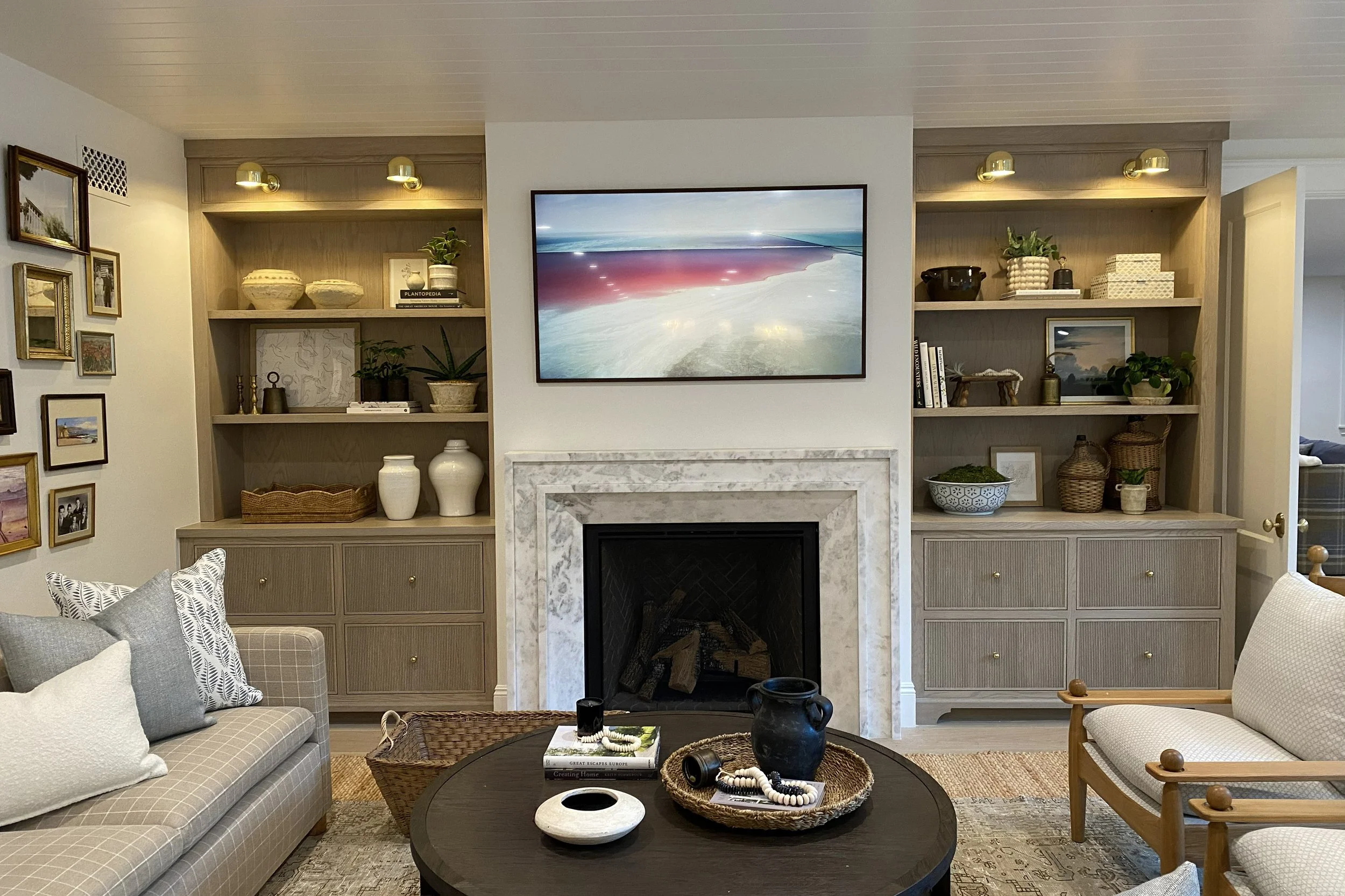 Living room with built-in wooden shelves, a marble fireplace, a wall-mounted TV, a gray checkered sofa adorned with pillows, a wicker coffee table with decorative objects, and framed artwork on the wall.