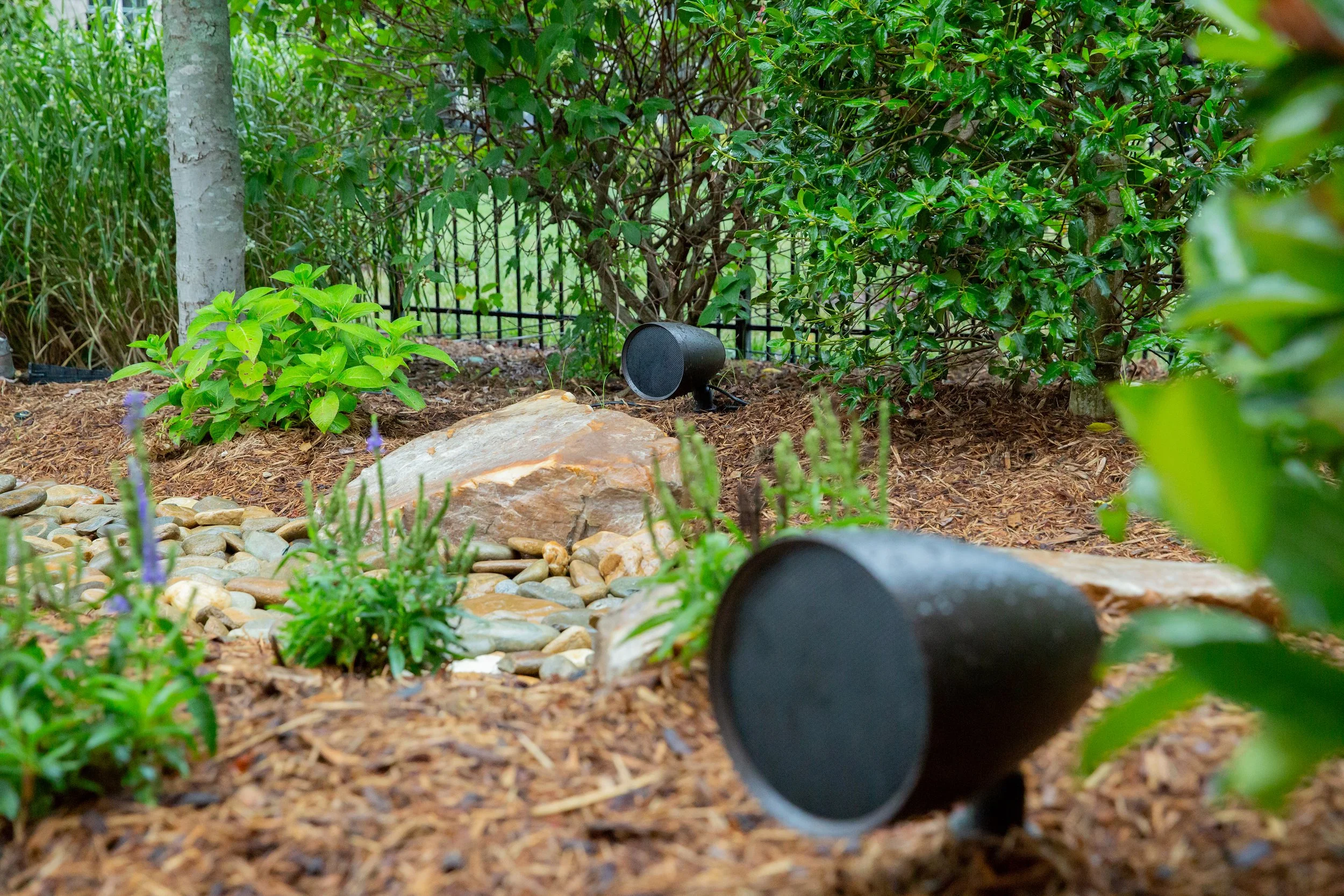Close-up of a landscaped garden with green shrubs, small plants, rocks, mulch, and black outdoor speaker, with a black metal fence in the background.