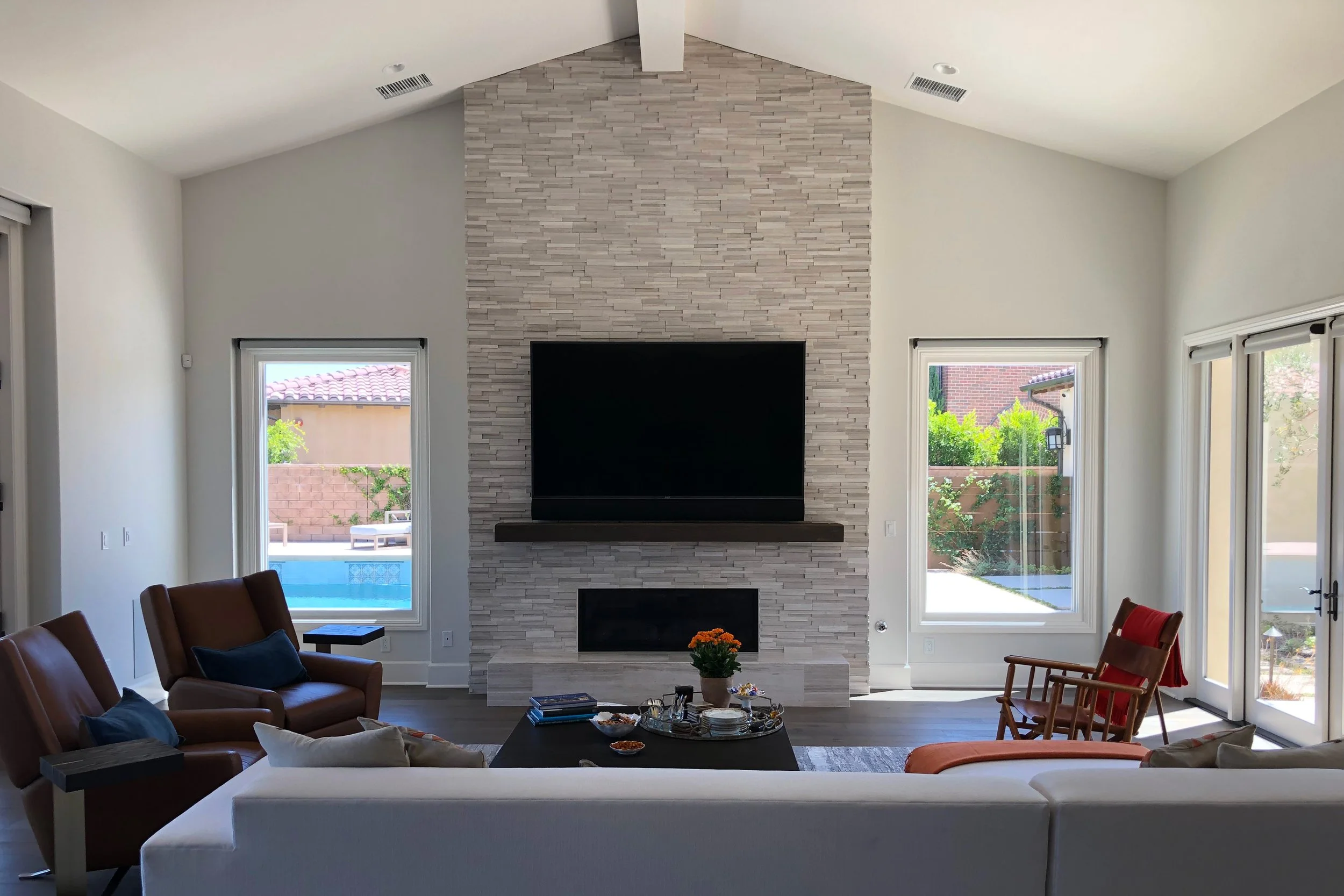Living room with a central white stone fireplace, large windows and sliding glass doors showing backyard, brown leather chairs with blue pillows, wooden chairs, and a coffee table with snacks and flowers.