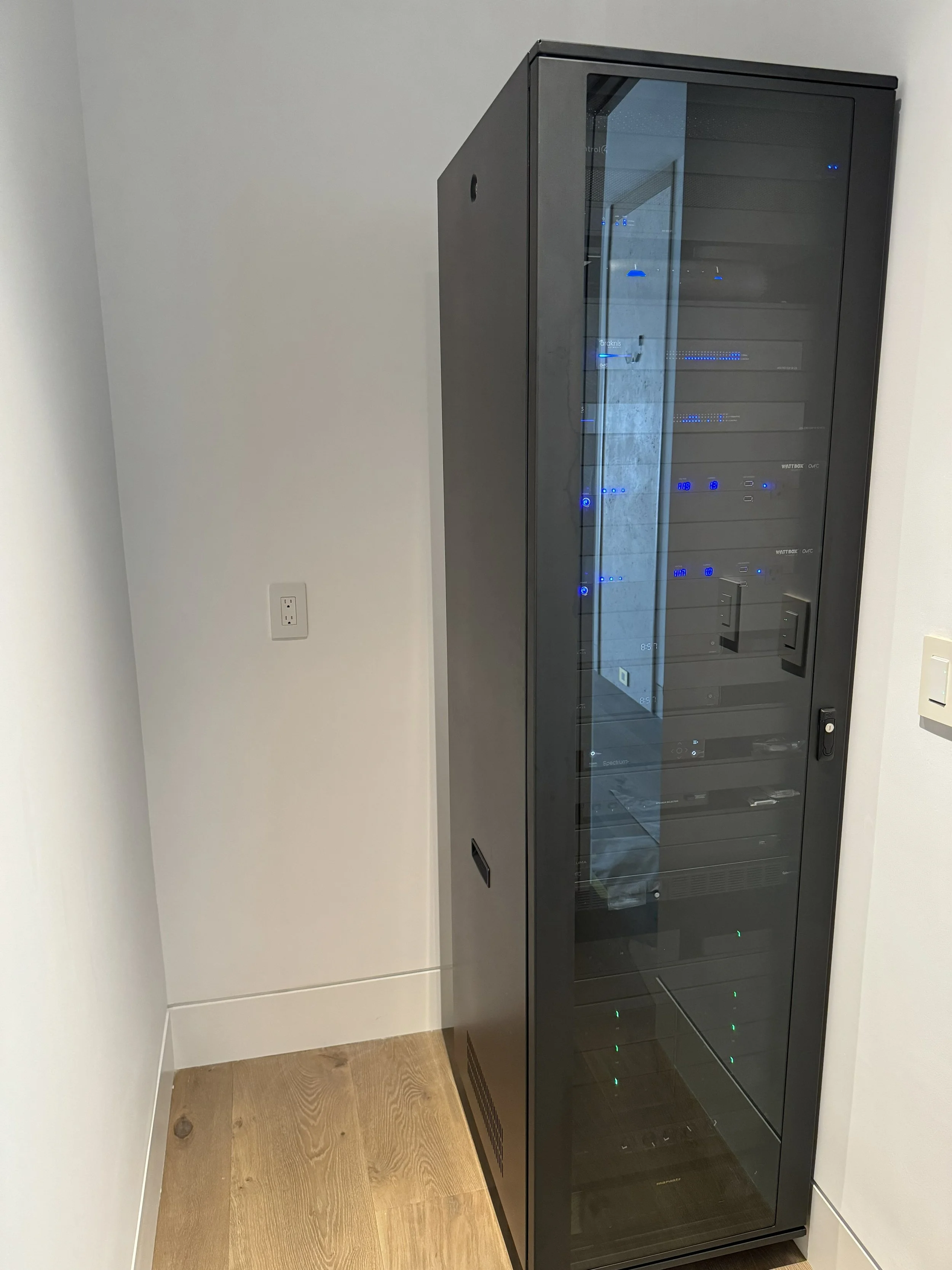 A black server rack with a glass front, situated in a white room with light wood flooring.