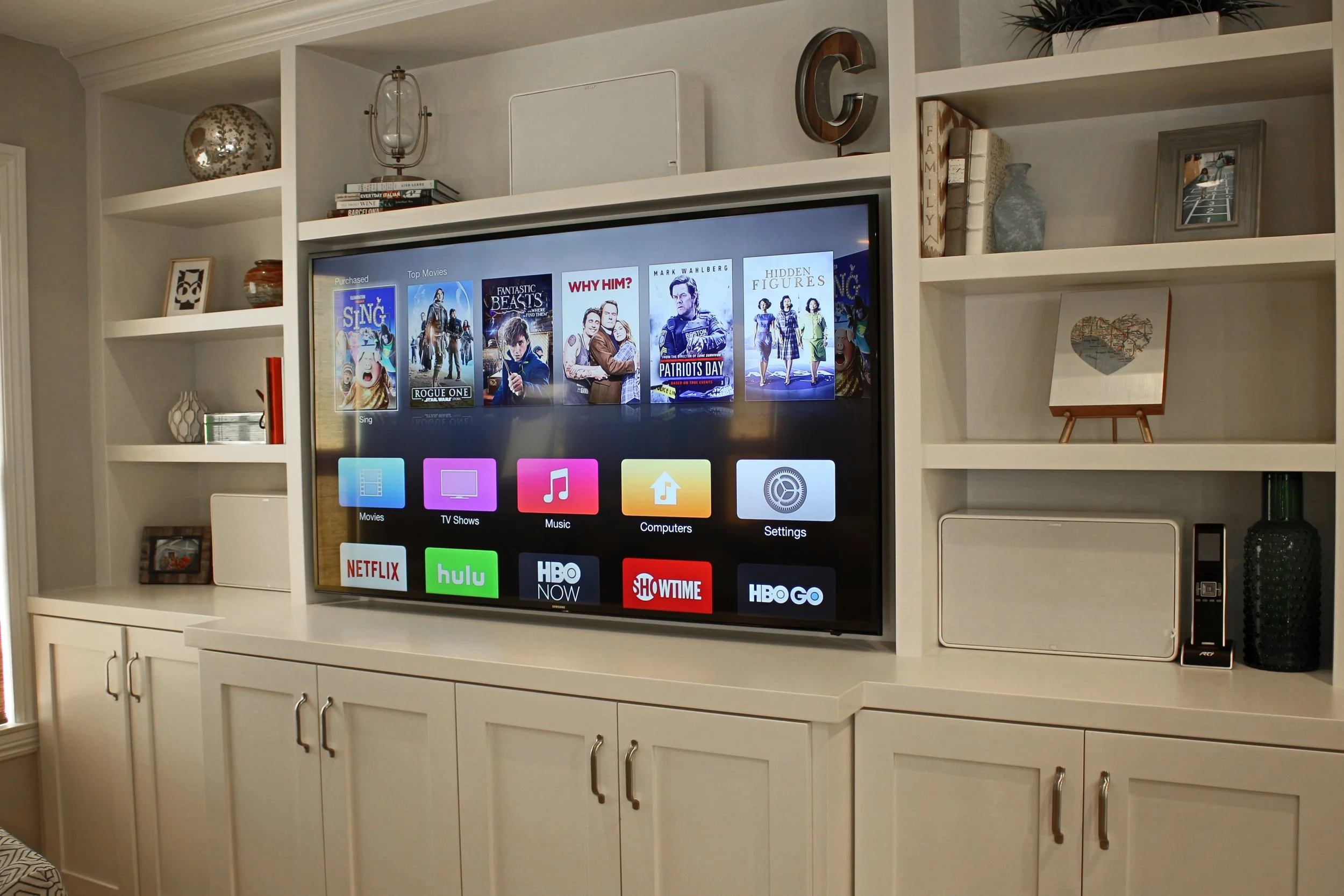 Living room entertainment center with a smart TV displaying various streaming apps, surrounded by shelves with decorative items, books, and picture frames.
