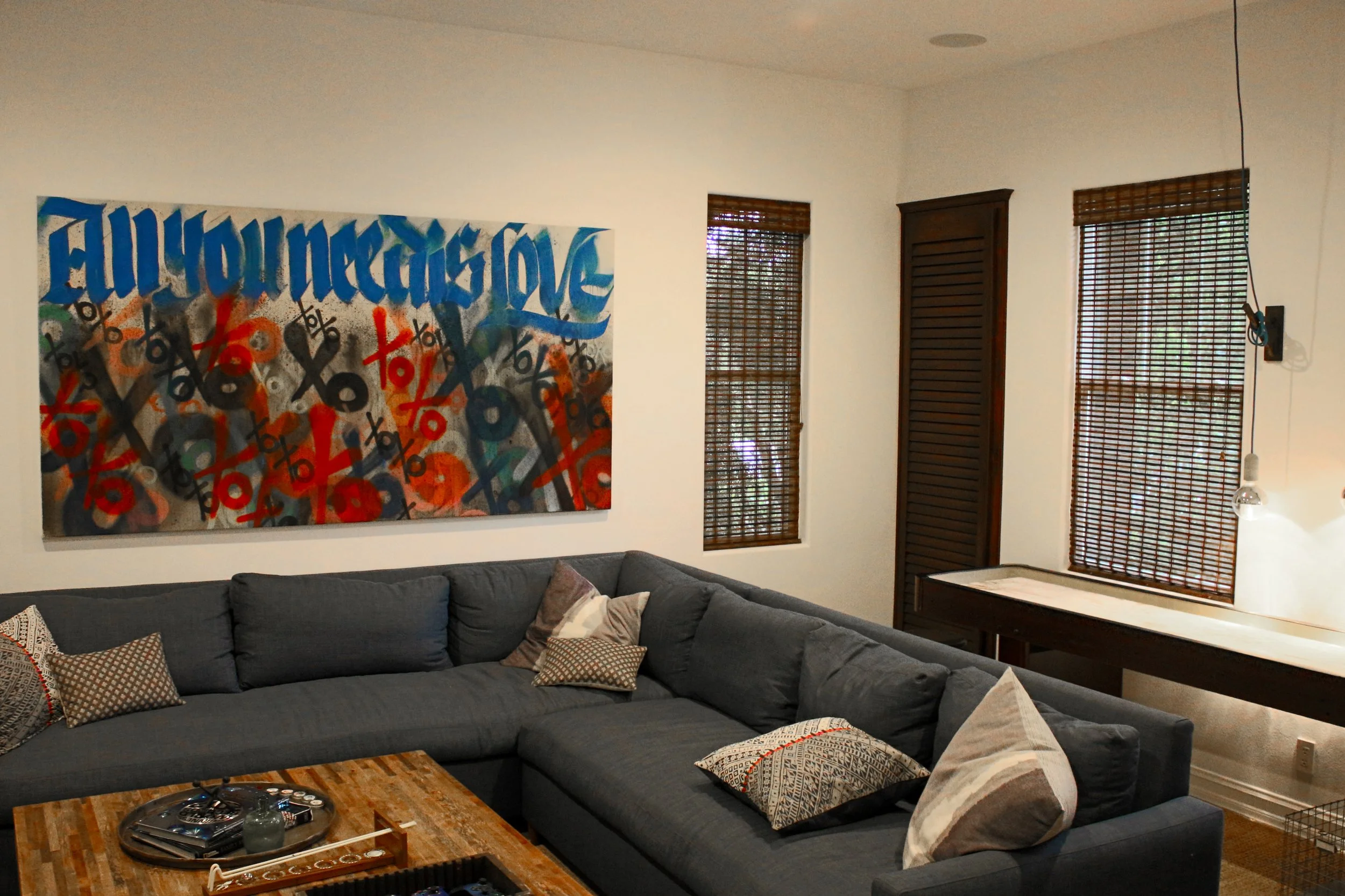A living room with a dark gray sectional sofa decorated with various patterned throw pillows, a wooden coffee table with a game and small items, a large colorful abstract painting on the wall, three windows with wooden blinds, and a closed wooden lou