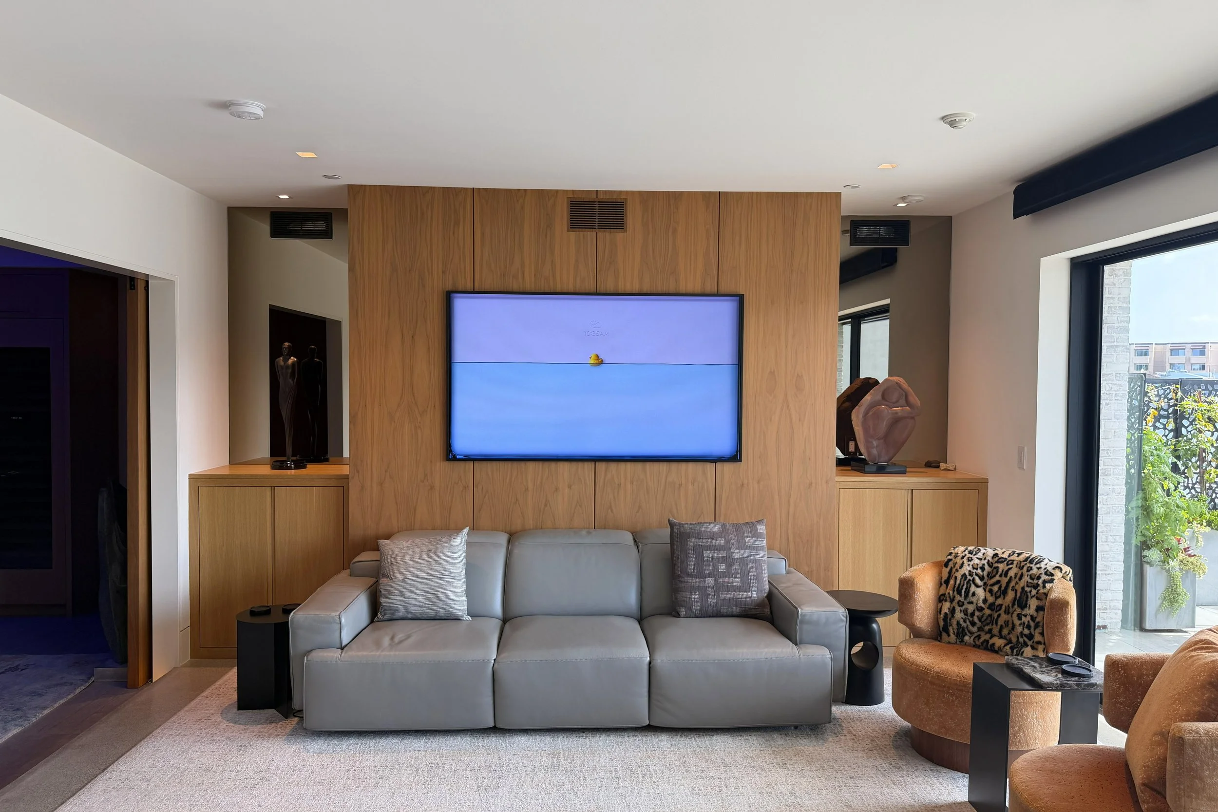 A modern living room with a flat-screen TV mounted on a wooden wall, a beige sofa with pillows, an orange patterned armchair, and large windows with black frames showing a building and greenery outside.