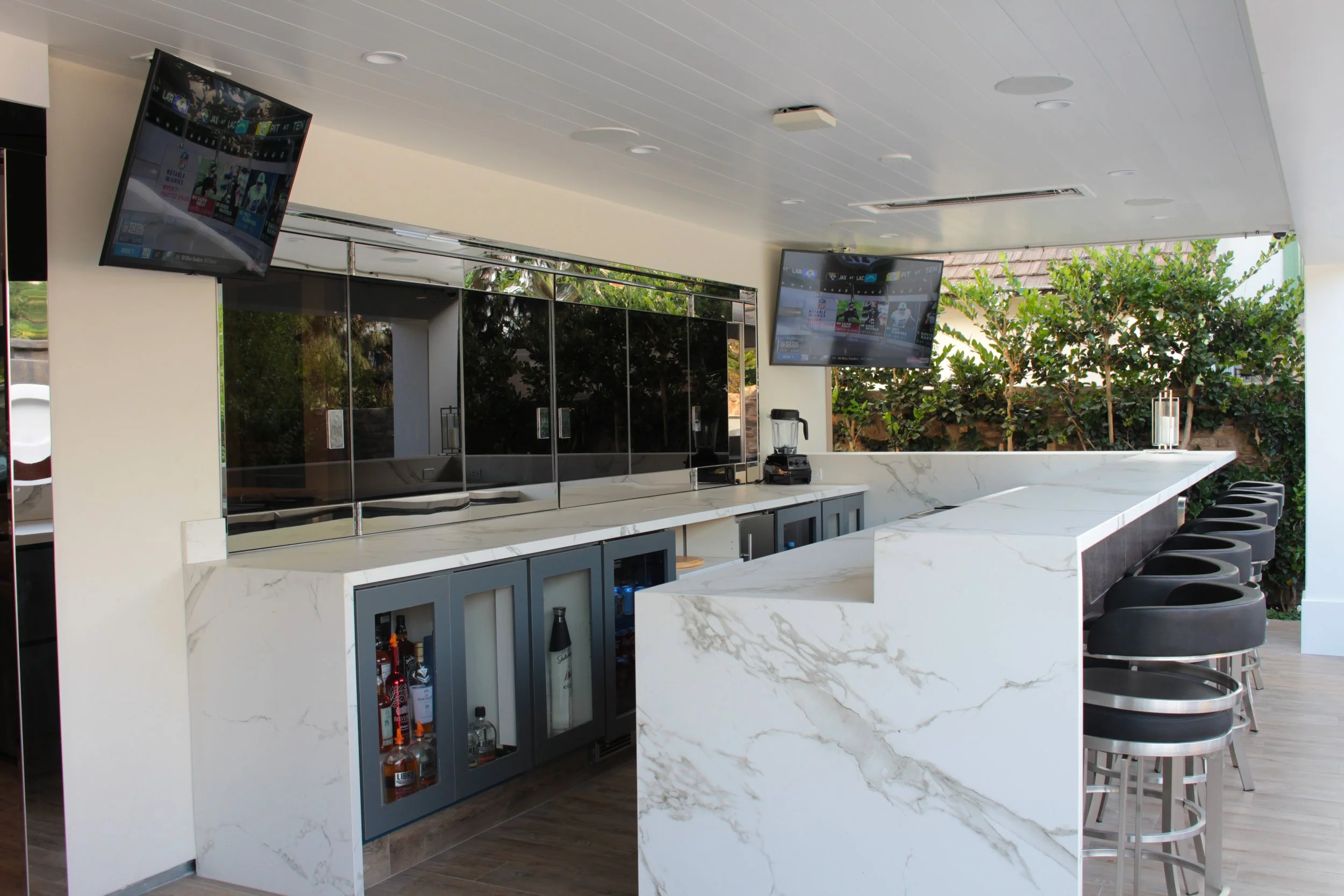 Modern outdoor bar area with a white marble countertop, bar stools, two wall-mounted televisions, a glass front refrigerator stocked with bottles, and lush green trees outside.