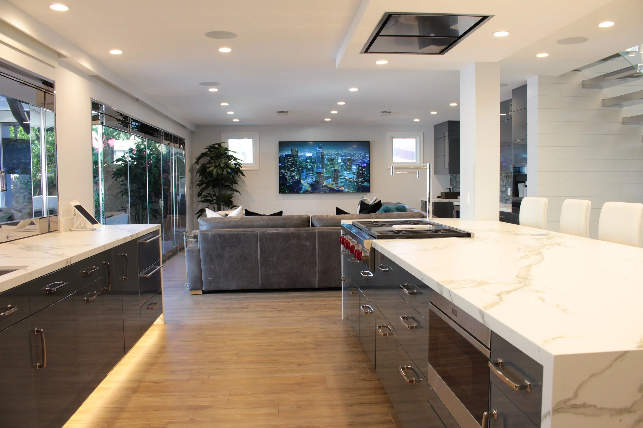 Open-concept modern kitchen and living room with white marble countertops, black cabinetry, hardwood floors, large window, wall-mounted TV showing a cityscape, and a staircase with metal railing.