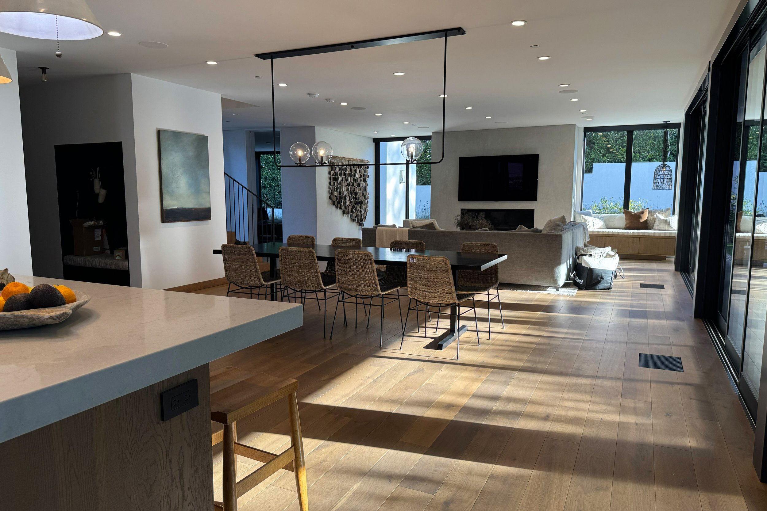 Open-concept living and dining area in a modern home with large windows, hardwood floors, a hallway, and a dining table with wicker chairs.