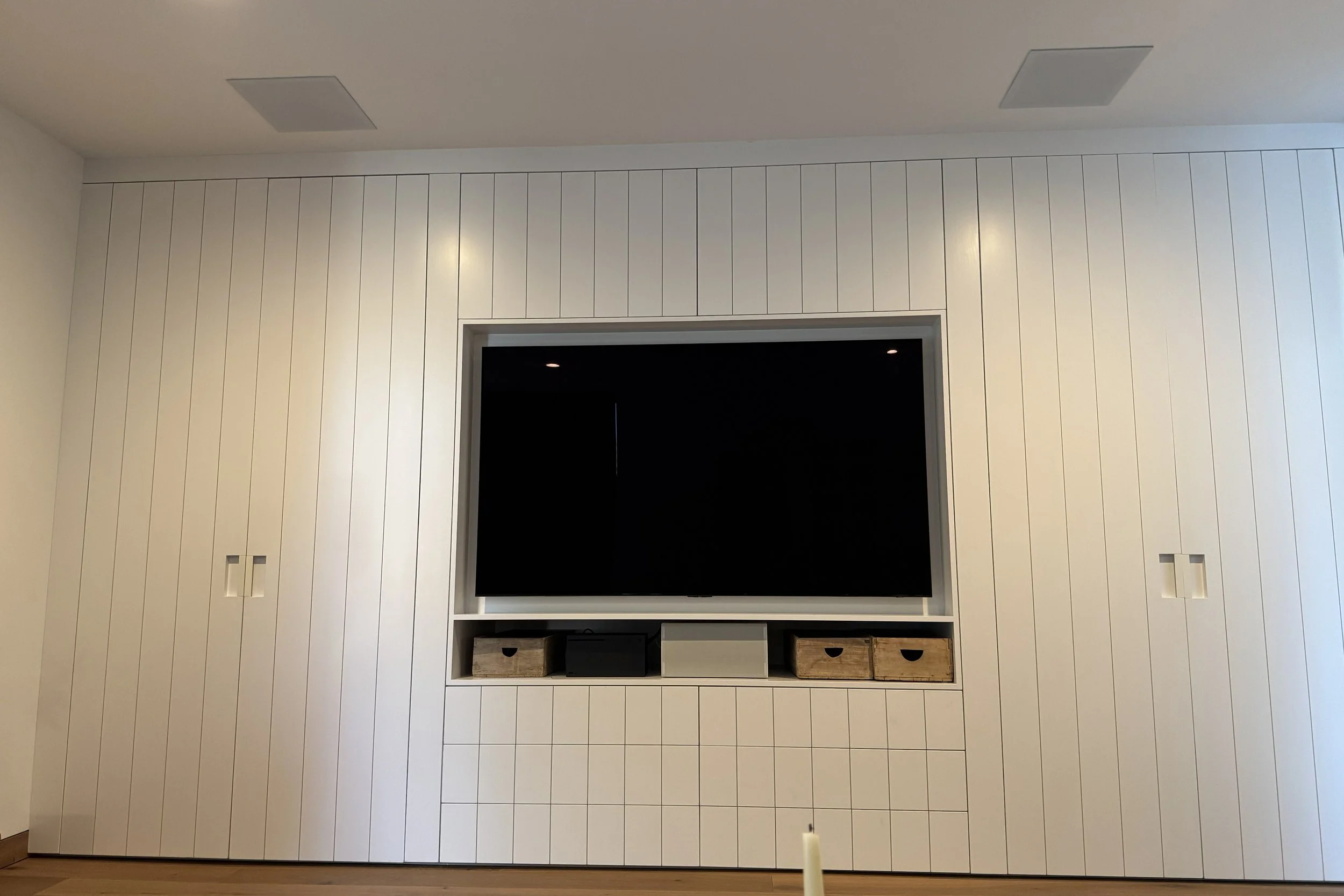 White entertainment center with a large flat-screen TV mounted on it, surrounded by vertical white paneling; includes shelves with baskets and electronic devices.