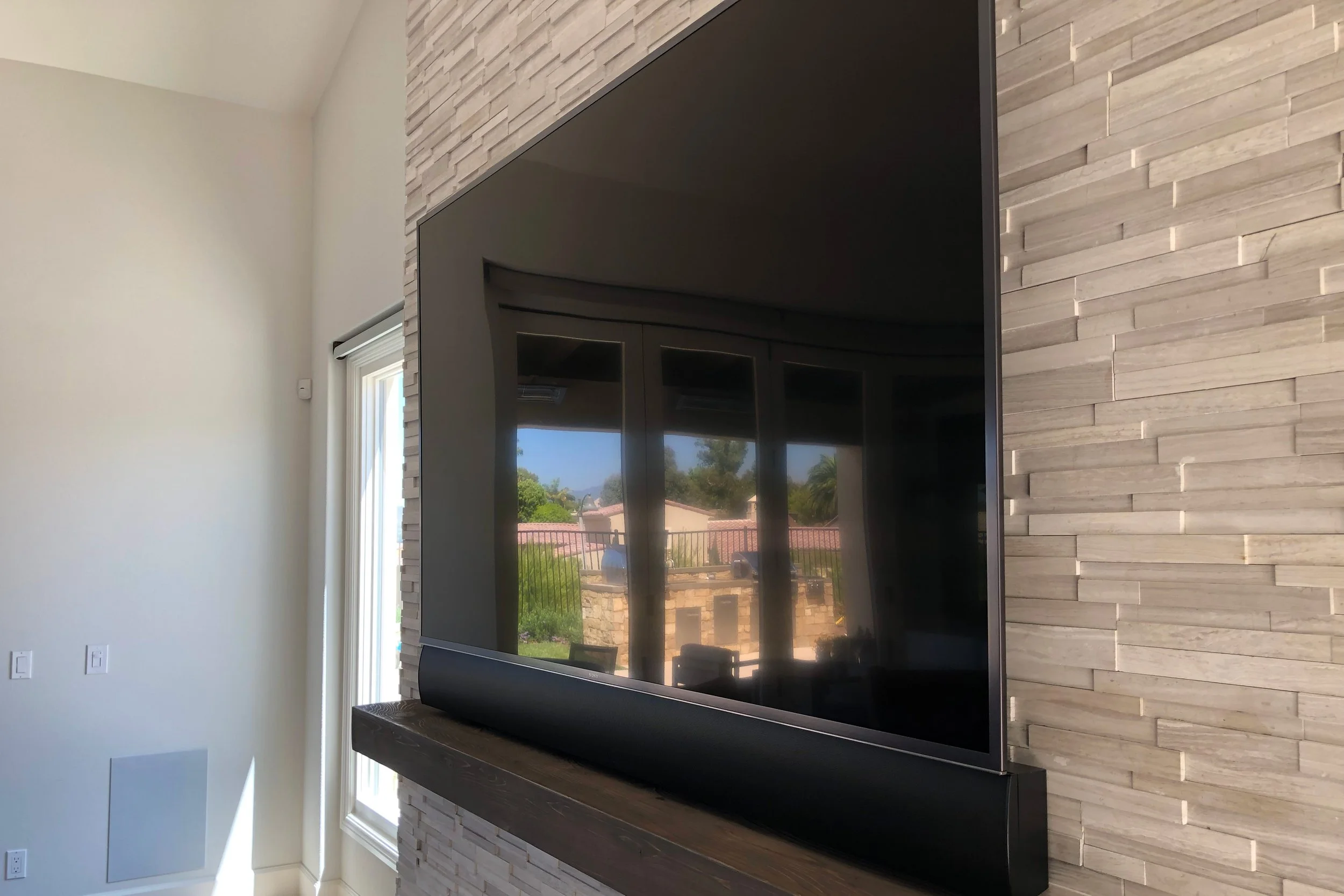 Flat-screen television mounted on a textured stone wall in a modern room. Large window with a view of trees and neighboring houses.