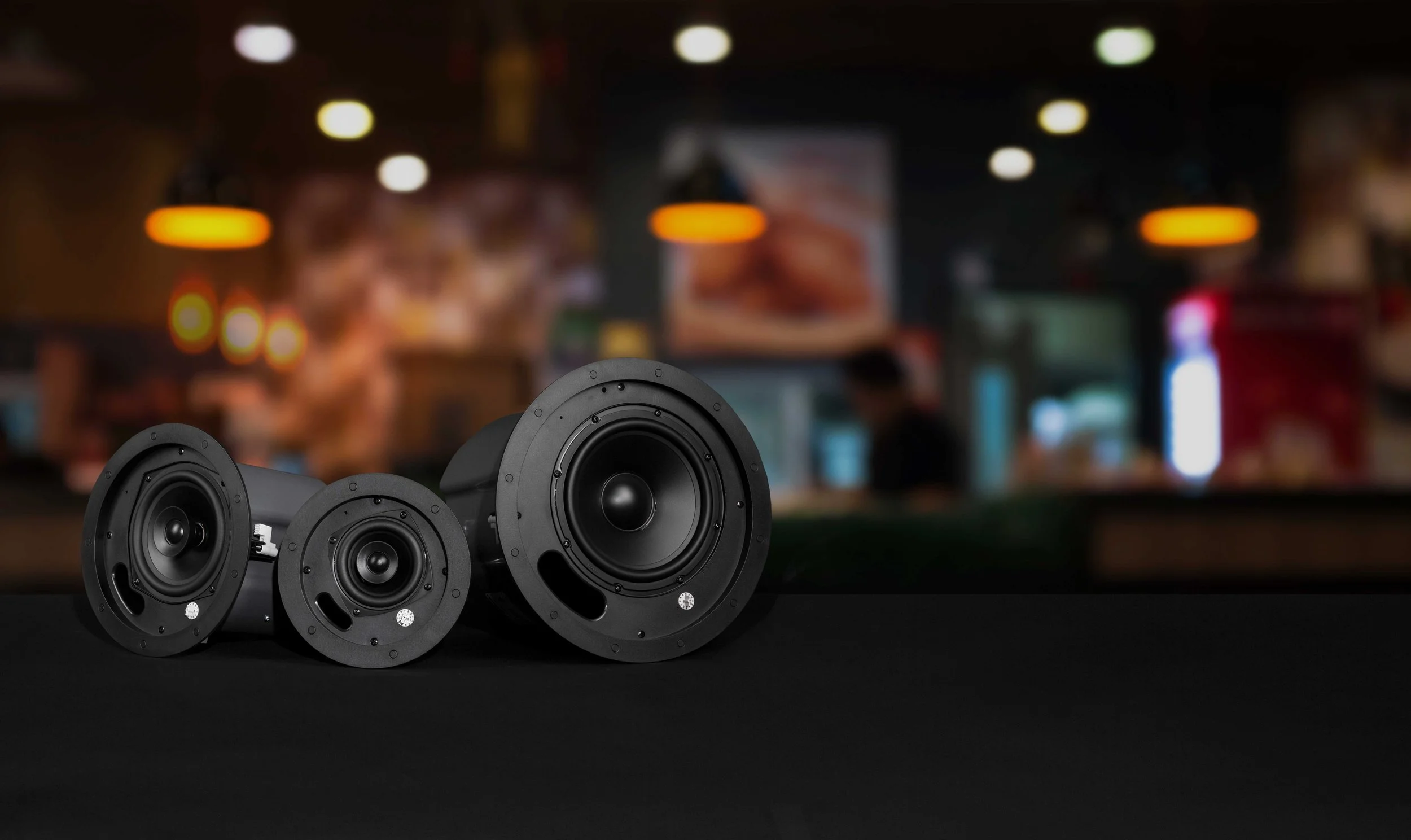 A set of black loudspeakers on a dark surface in a dimly lit indoor setting, with blurred background lights.