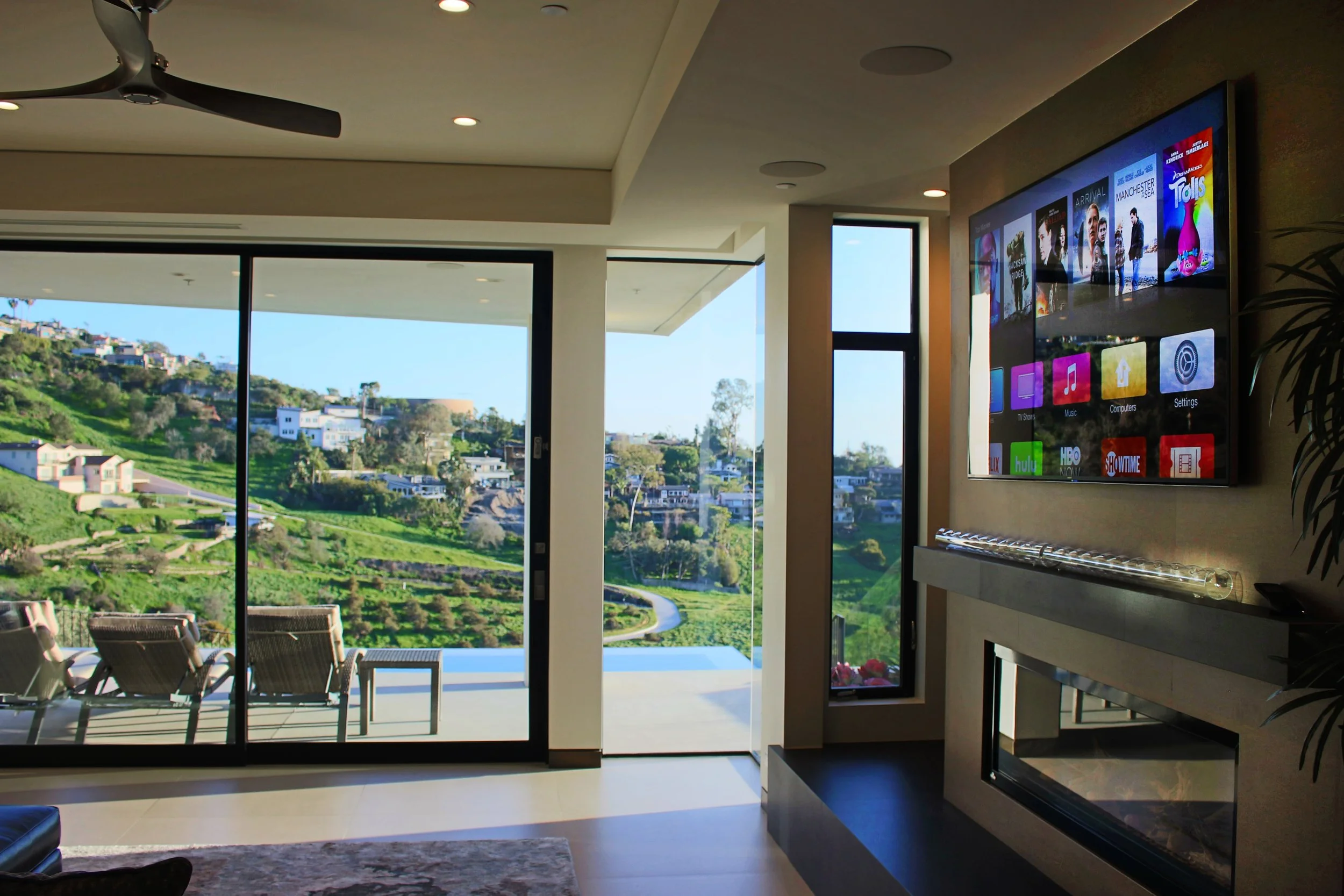 Modern living room with large glass sliding doors leading to a patio with chairs overlooking green hills and houses.