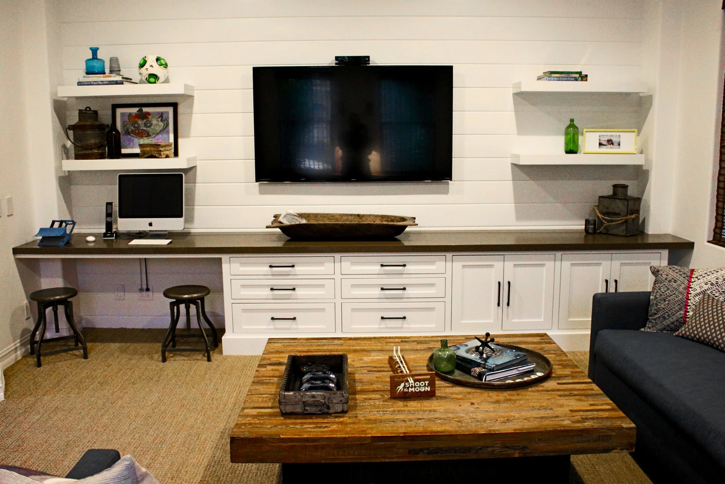 Living room with a white shiplap wall, mounted flat-screen TV, two white shelves with decorative items, a wooden console table, and a dark sofa with a patterned pillow. There are two small stools in front of the console.