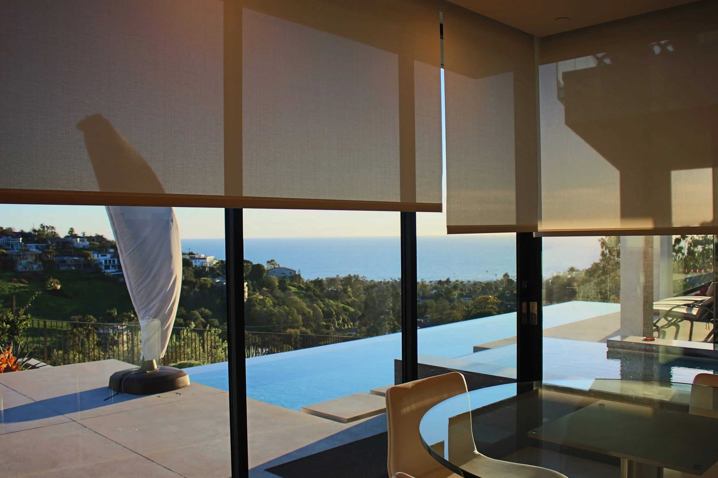 Interior view of a modern home with a large sliding glass door opening to a scenic outdoor area. Through the door, there is a backyard with a swimming pool, lush greenery, coastal houses, and the ocean in the distance under a clear sky. The scene is 