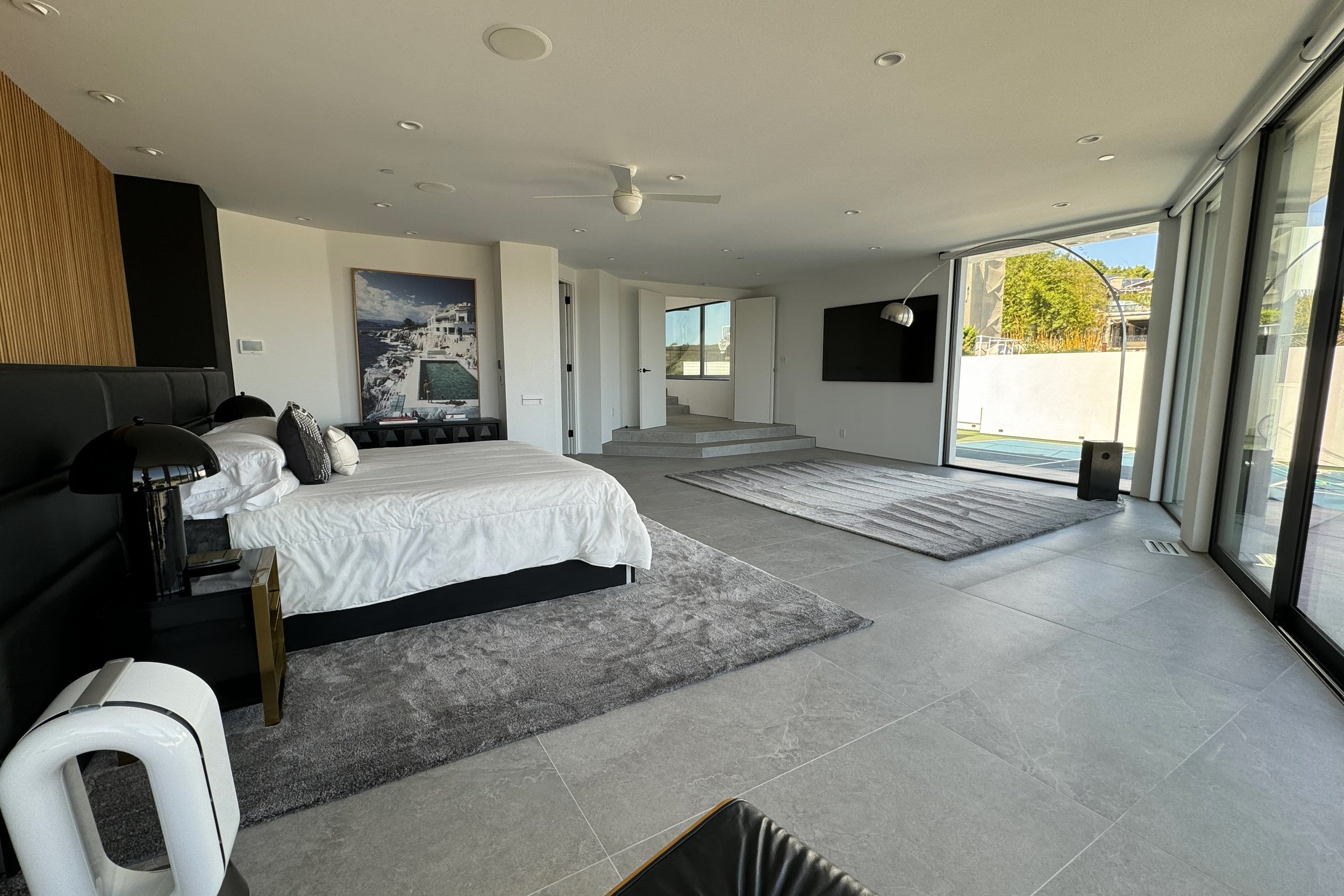 Modern bedroom with large sliding glass doors leading to an outdoor space, contemporary decor, gray tiled flooring, a bed with black headboard, side lamps, and wall art.