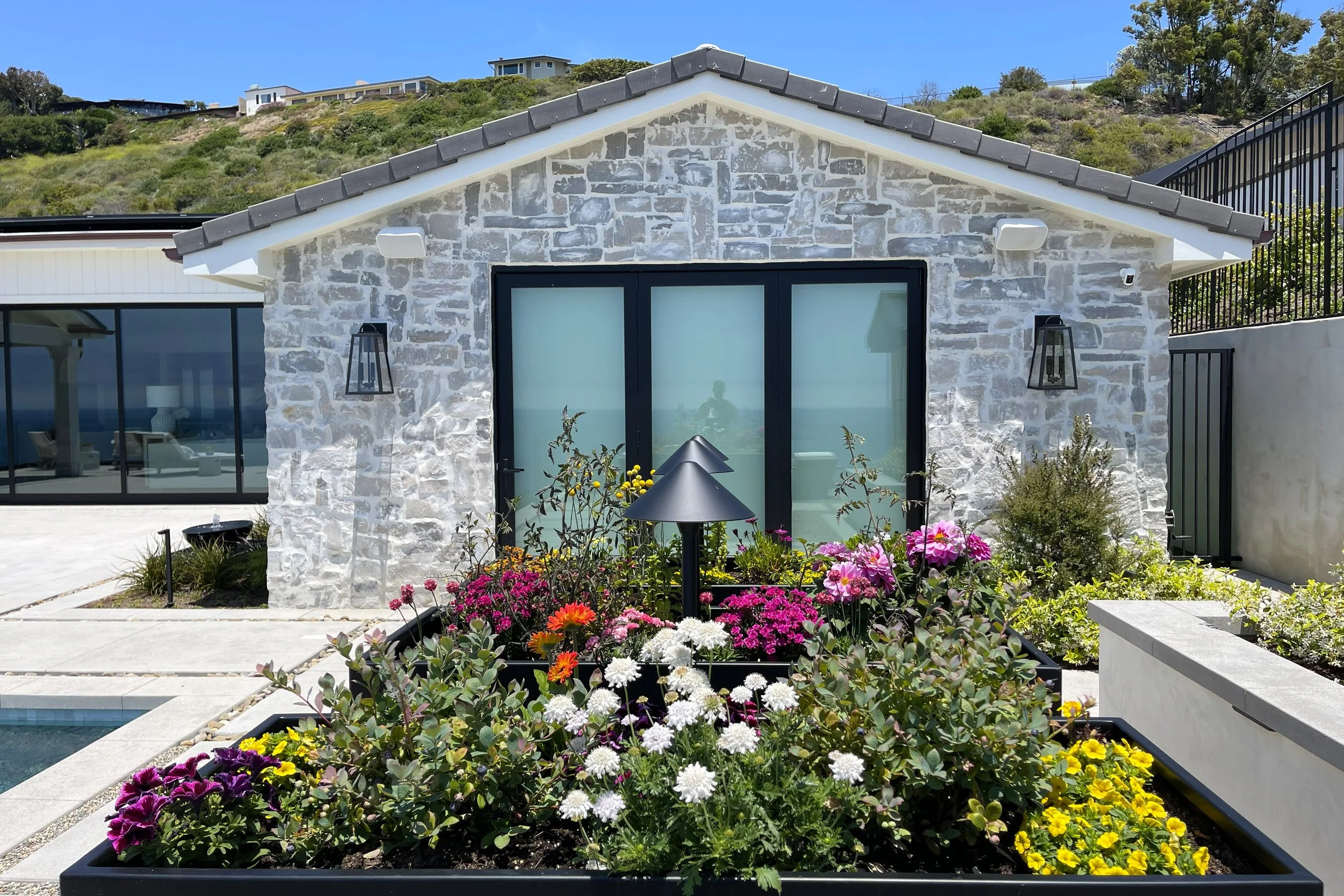 A modern house with a stone facade, black-framed sliding glass door, and outdoor lighting, set against a hillside with green vegetation and other houses in the background, with a front garden filled with colorful flowers.