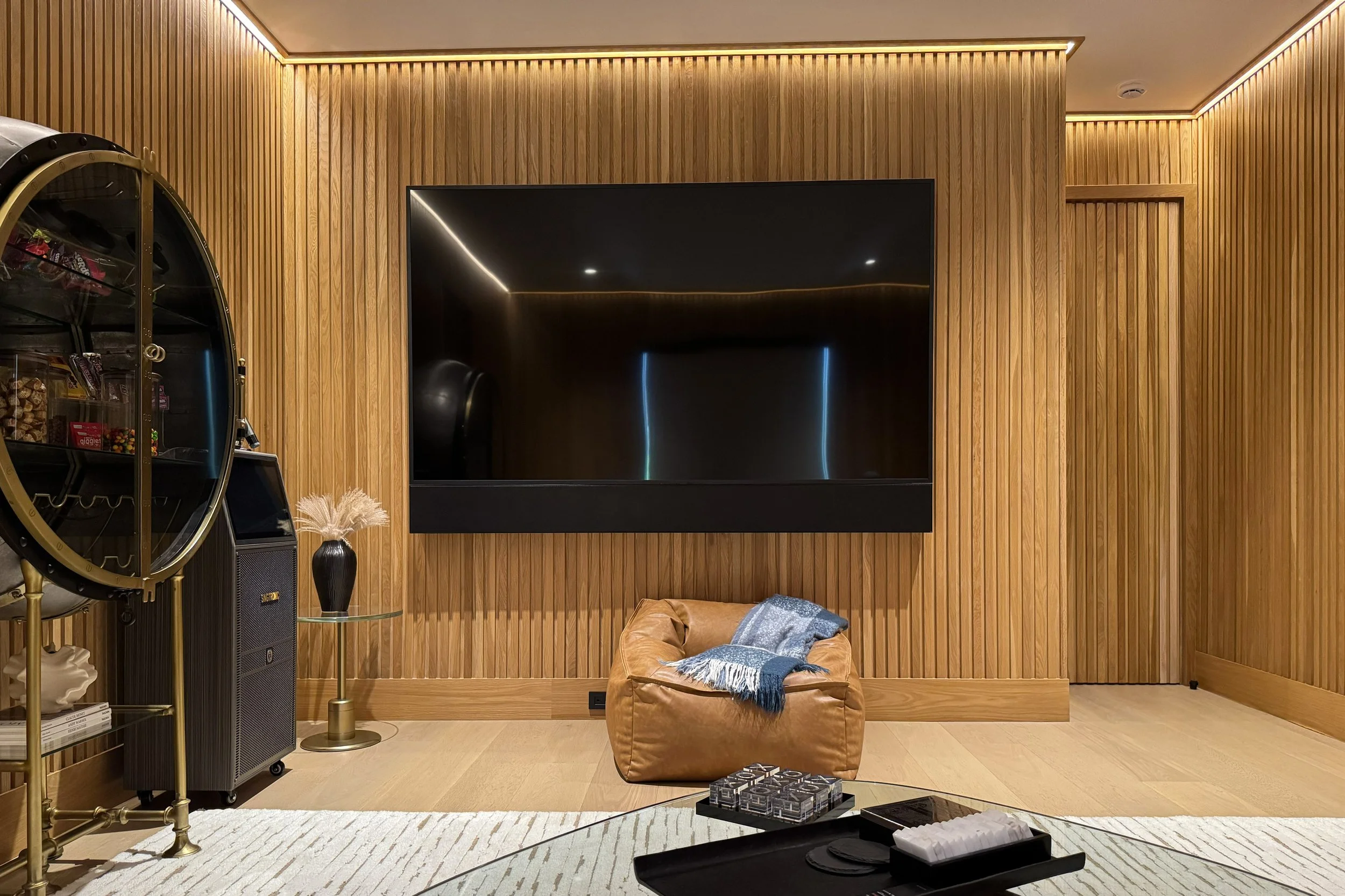 Living room with vertical wooden paneling, a large black flat-screen TV mounted on the wall, a brown leather ottoman with a blue blanket, a small table with a decorative vase, a popcorn vending machine, and a rug on light wood flooring.