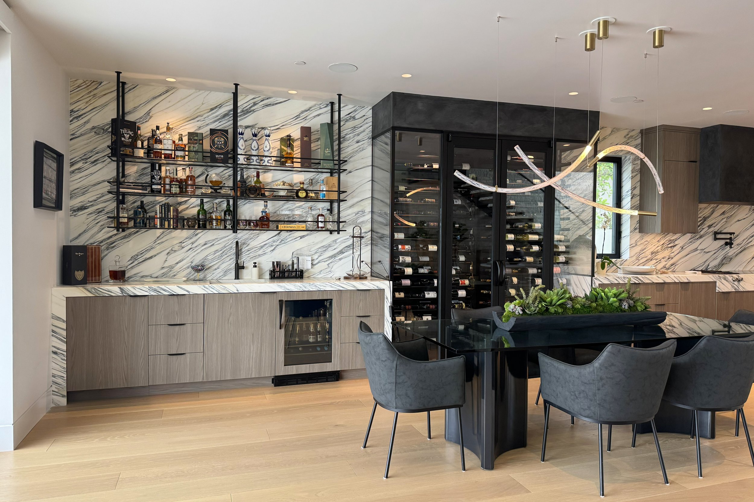 Modern kitchen with marbled walls, wine refrigerator, black shelving with liquor bottles, a black dining table with gray chairs, and green plants in a black planter.