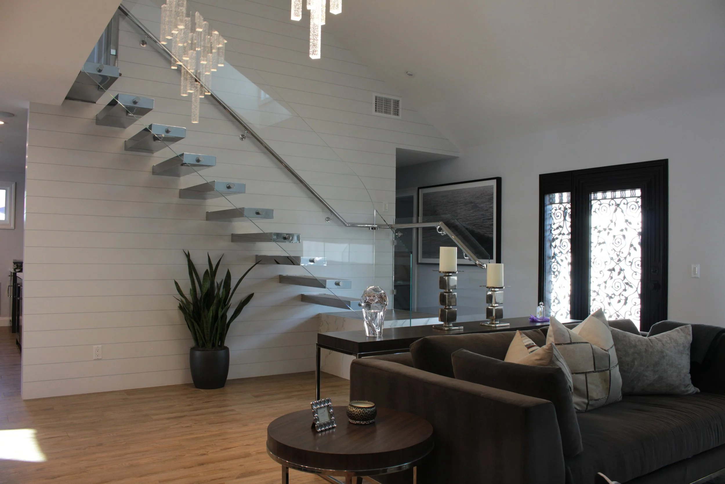 Modern living room with a staircase featuring glass railings, a large potted plant, a gray sectional sofa with pillows, a dark wooden coffee table, candles, and decorative objects. Large windows with ornate ironwork allow natural light into the space