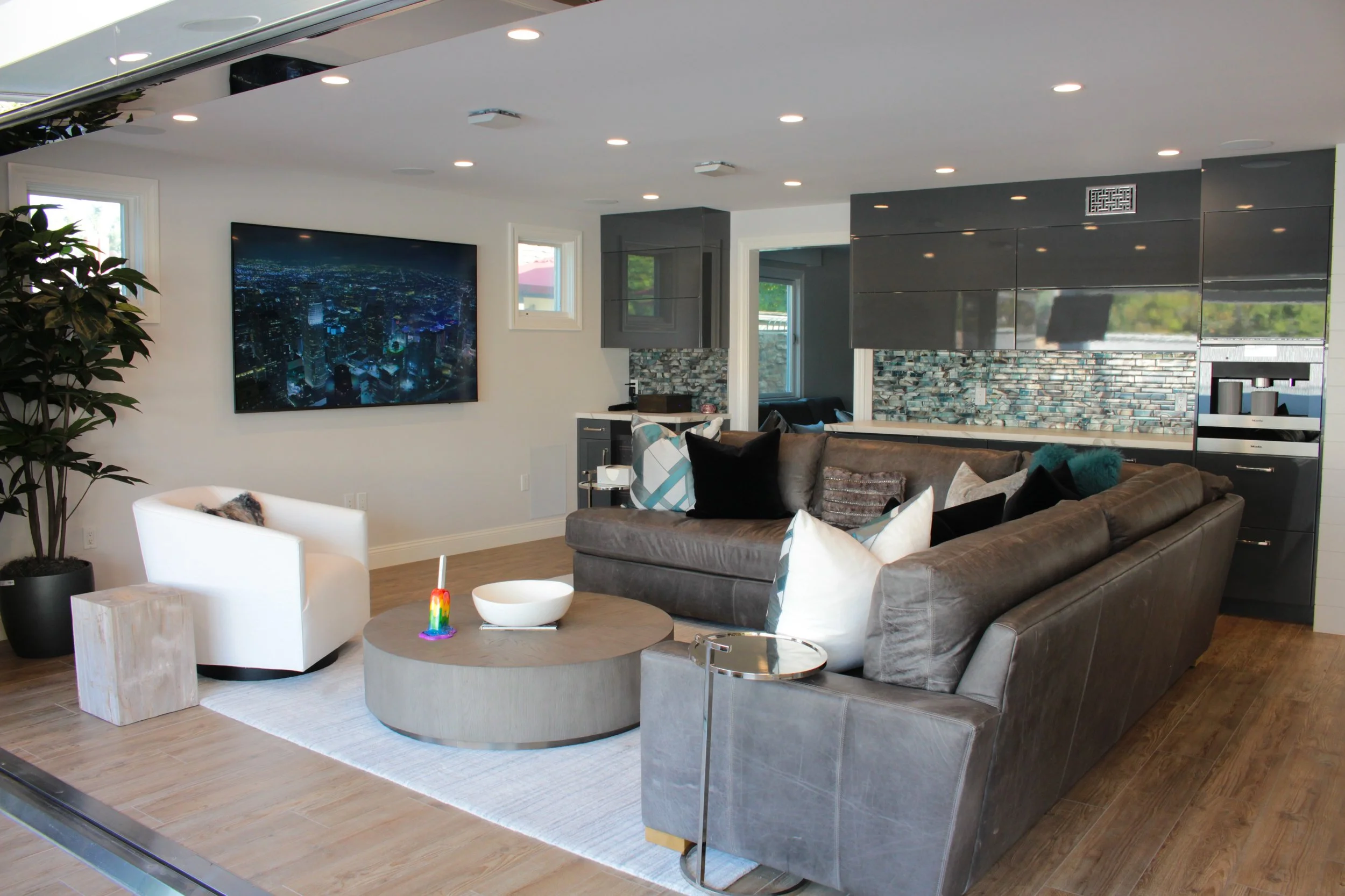 Modern living room with a large gray sectional sofa, a white armchair, and two round coffee tables. There is a large flat-screen TV displaying a cityscape at night, a potted plant, and decorative pillows on the sofa. The room has recessed lighting an