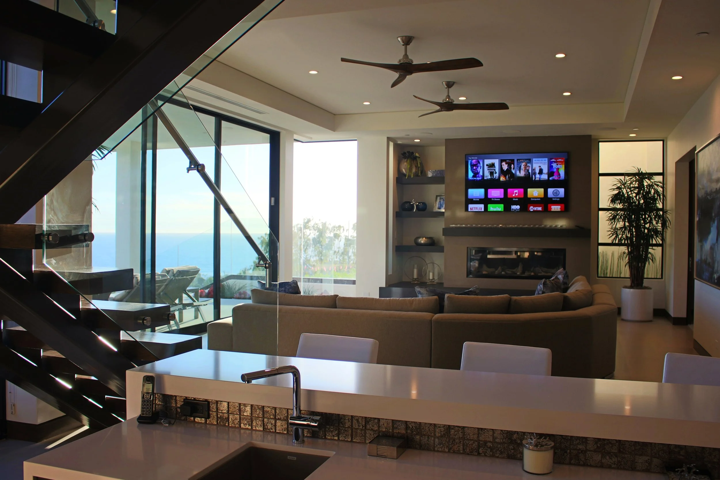 Modern living room with large windows showing ocean view, beige sectional sofa, wall-mounted TV, and ceiling fans.