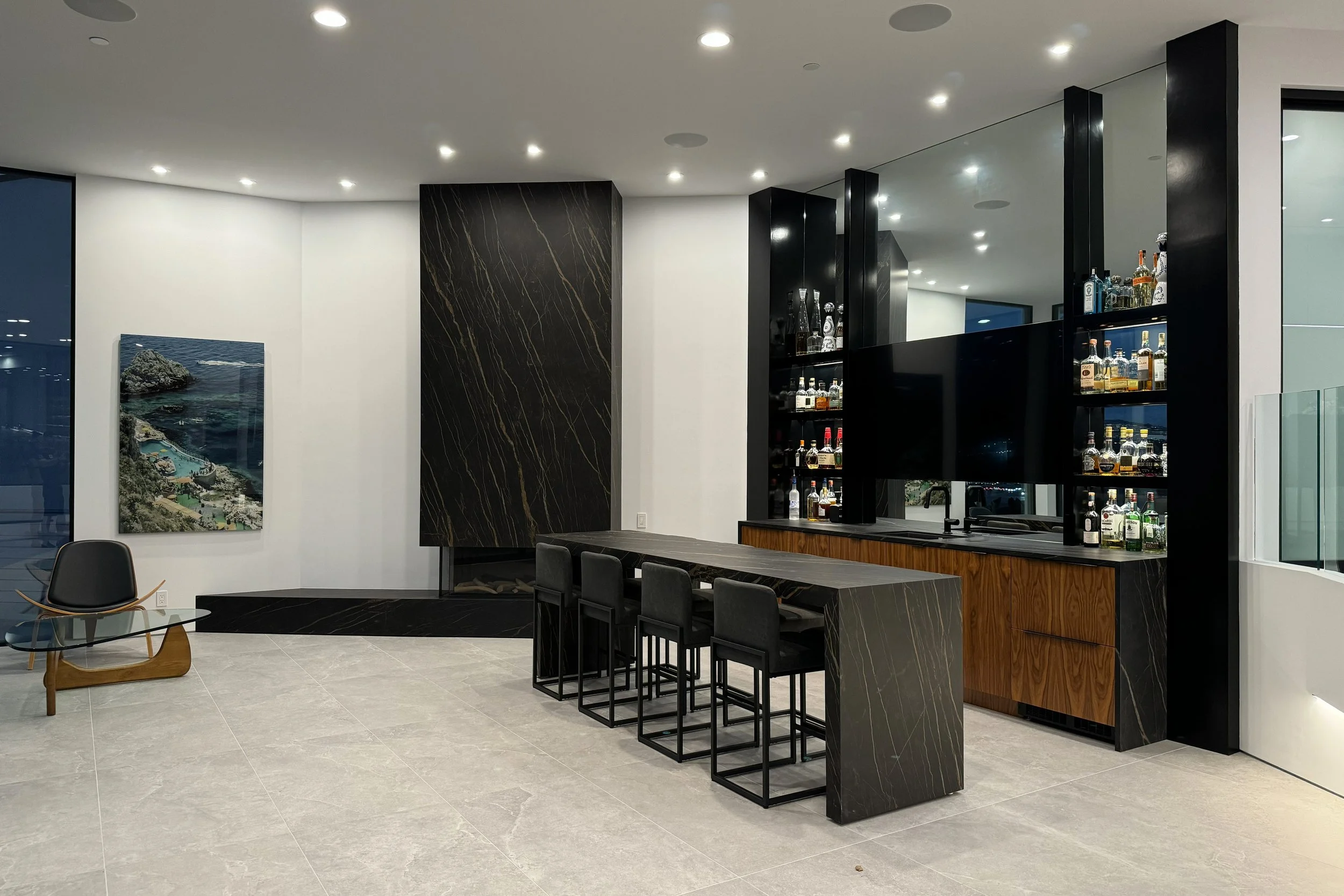 Modern indoor space featuring a bar area with black shelves stocked with liquor bottles, black and wood accents, and a seating area with a chair and a painting of a coastline.