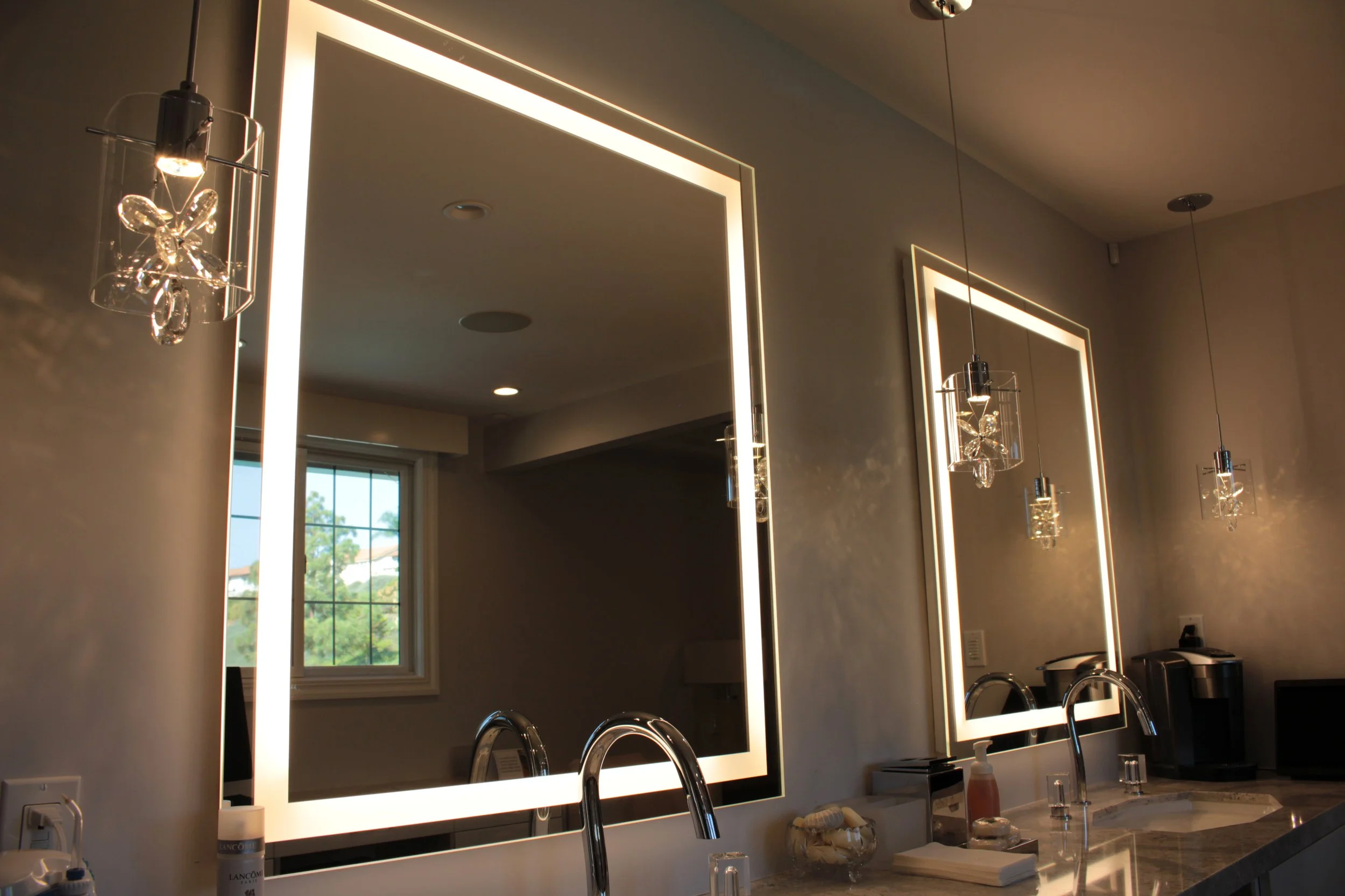 Modern bathroom with illuminated mirrors, sinks, faucets, skincare products, and a window with a view of trees outside.