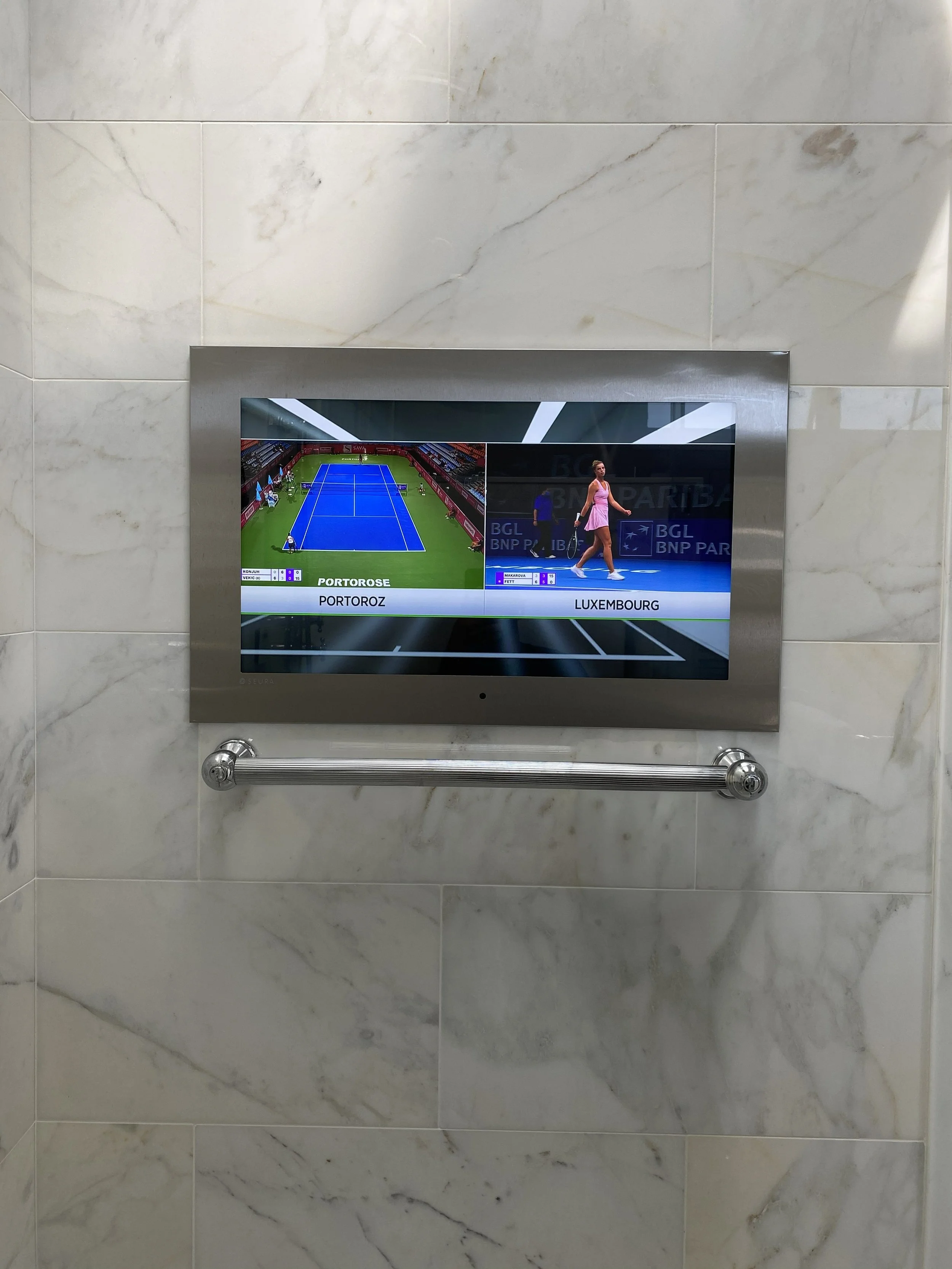 A flat screen display in a marble wall showing two live tennis matches, with players on the court and score information.
