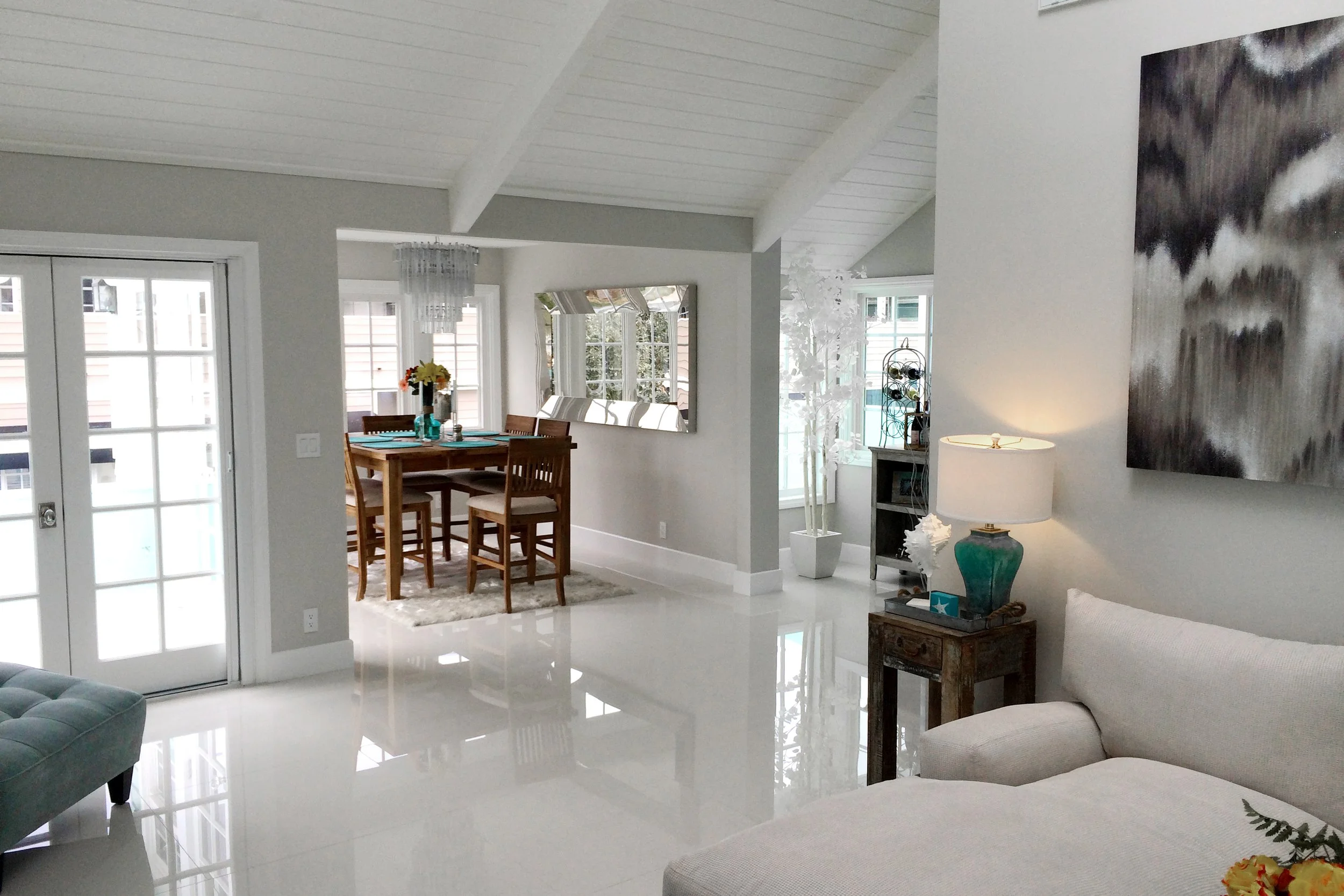 Bright, modern living and dining area with white tiled floors, large windows, a wooden dining table with chairs, a large mirror on the wall, a white decorative tree, and a colorful abstract painting.