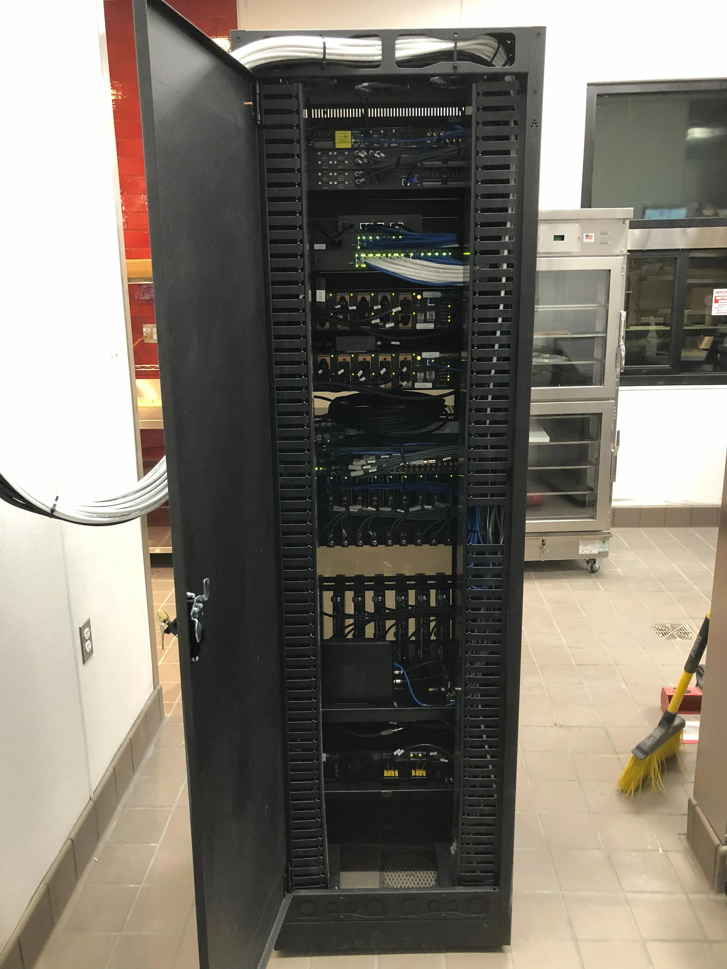 A tall black network server rack with its door open, filled with various networking equipment and cables, in a utility room.