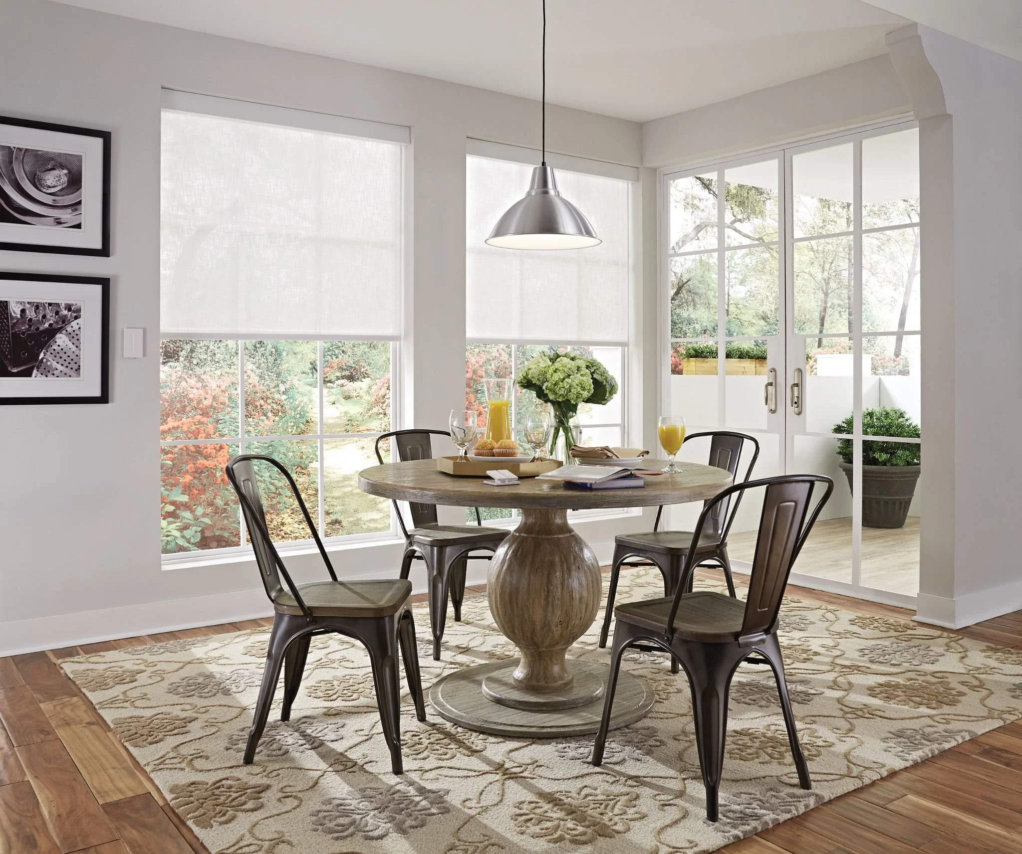 A dining area with a round wooden table, six metal chairs, a large window with blinds, a hanging silver pendant light, a floral centerpiece, and a patterned rug on wooden floorboards.