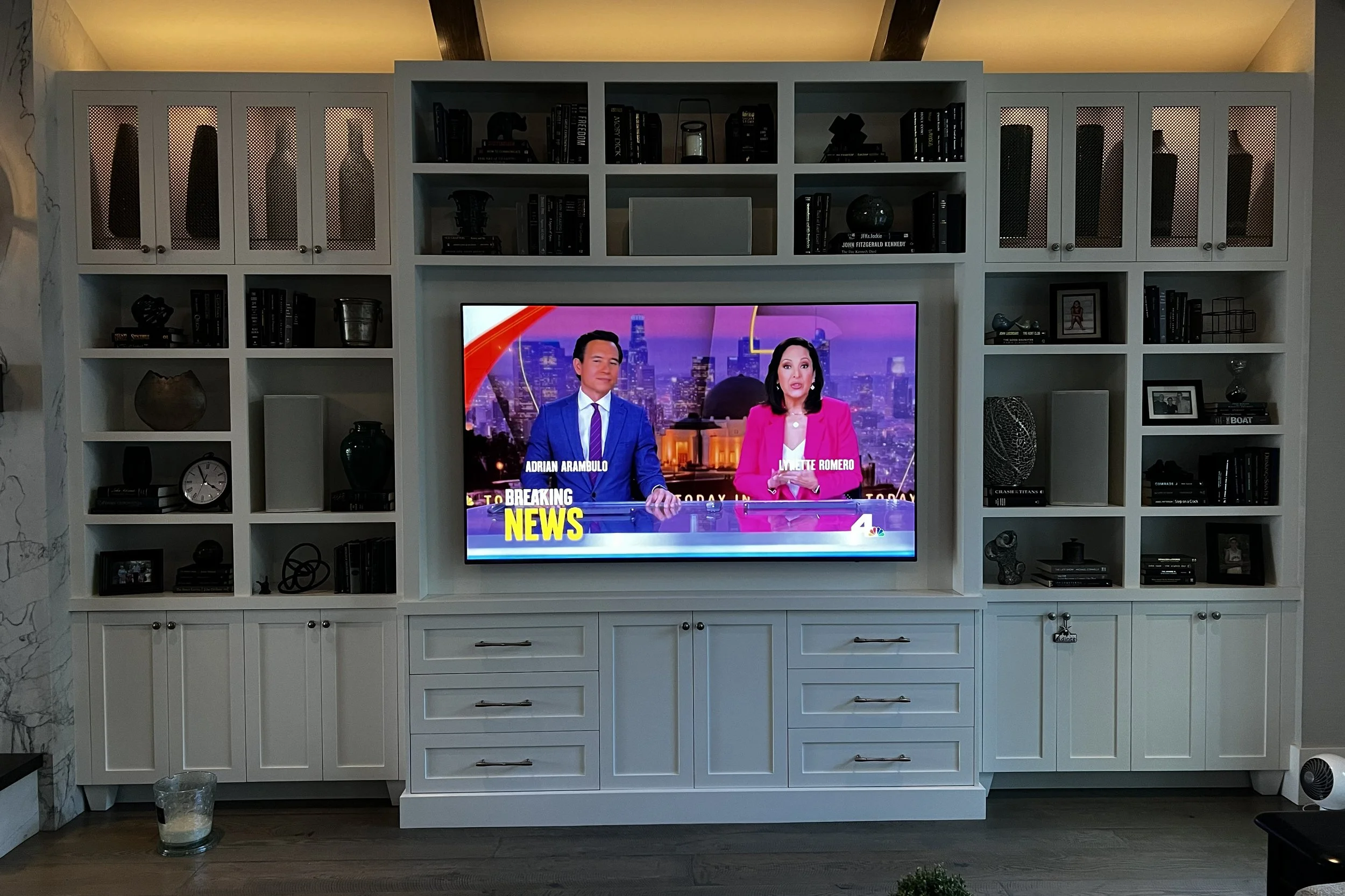 Living room with white built-in shelving unit containing books, decorative items, and framed photos. A flat-screen TV displays a news broadcast with a male and female news anchor. A glass of water is on the floor in front of the shelf, and a small po