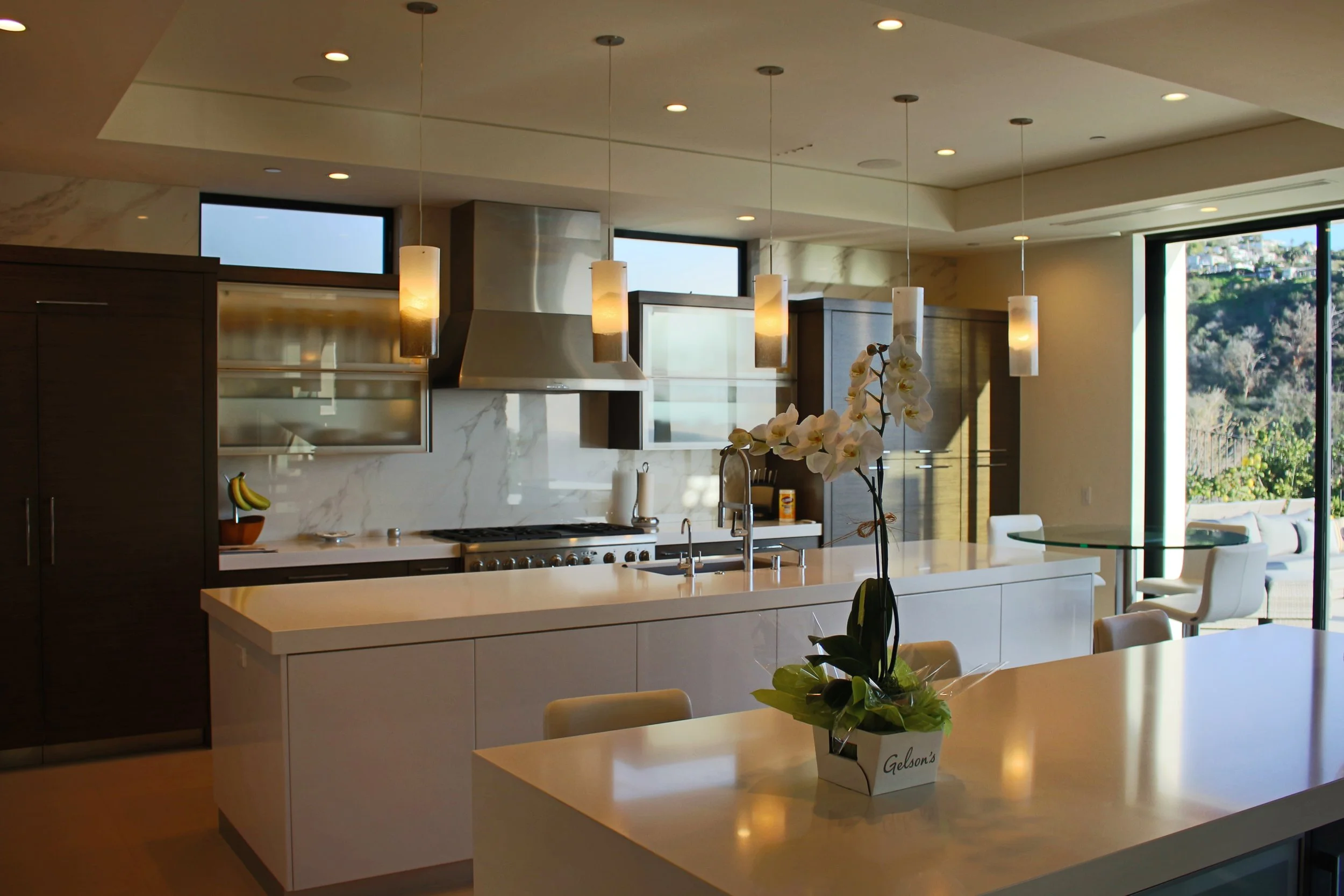 Modern kitchen with white countertops, wooden cabinets, stainless steel appliances, pendant lighting, and sliding glass doors revealing outdoor landscape.