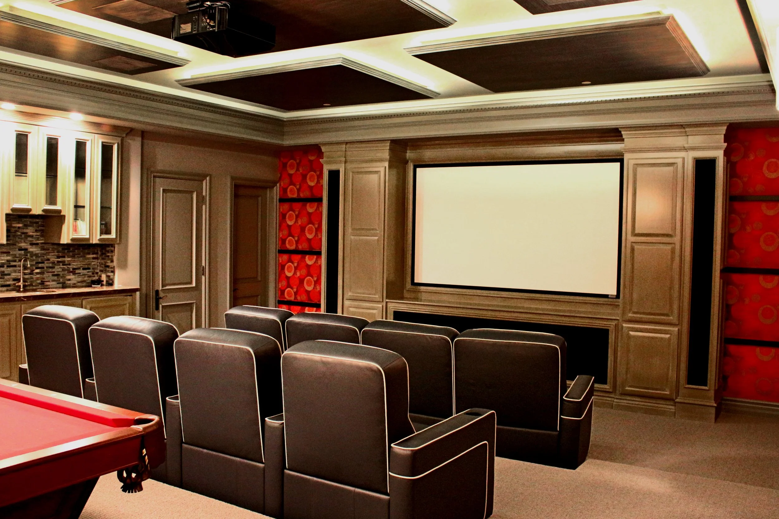 Home theater room with a large projection screen, black theater chairs, a pool table, and decorative wood paneling on the ceiling.