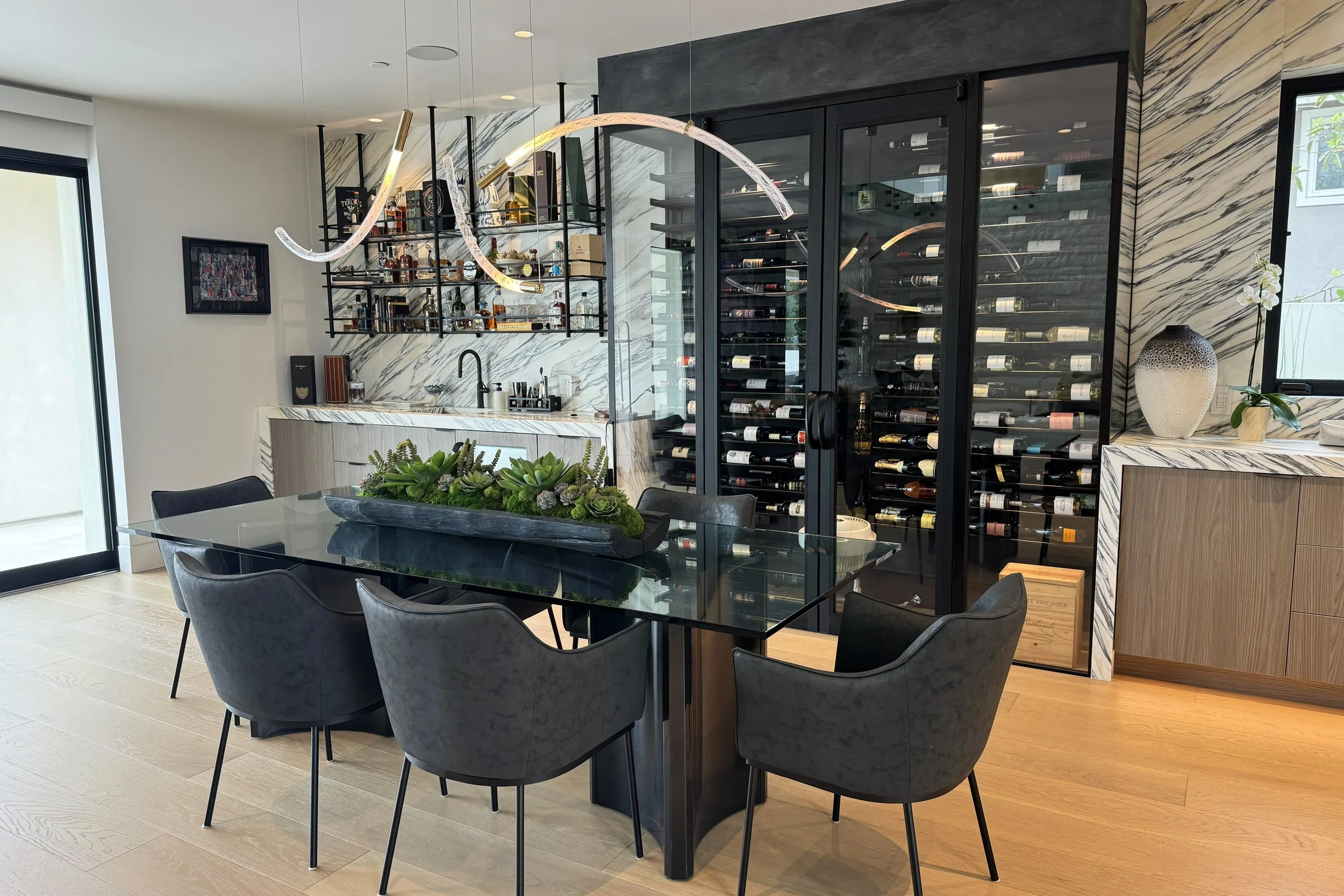Modern home bar area with a glass table and black chairs, featuring a wine refrigerator and decorative plants.