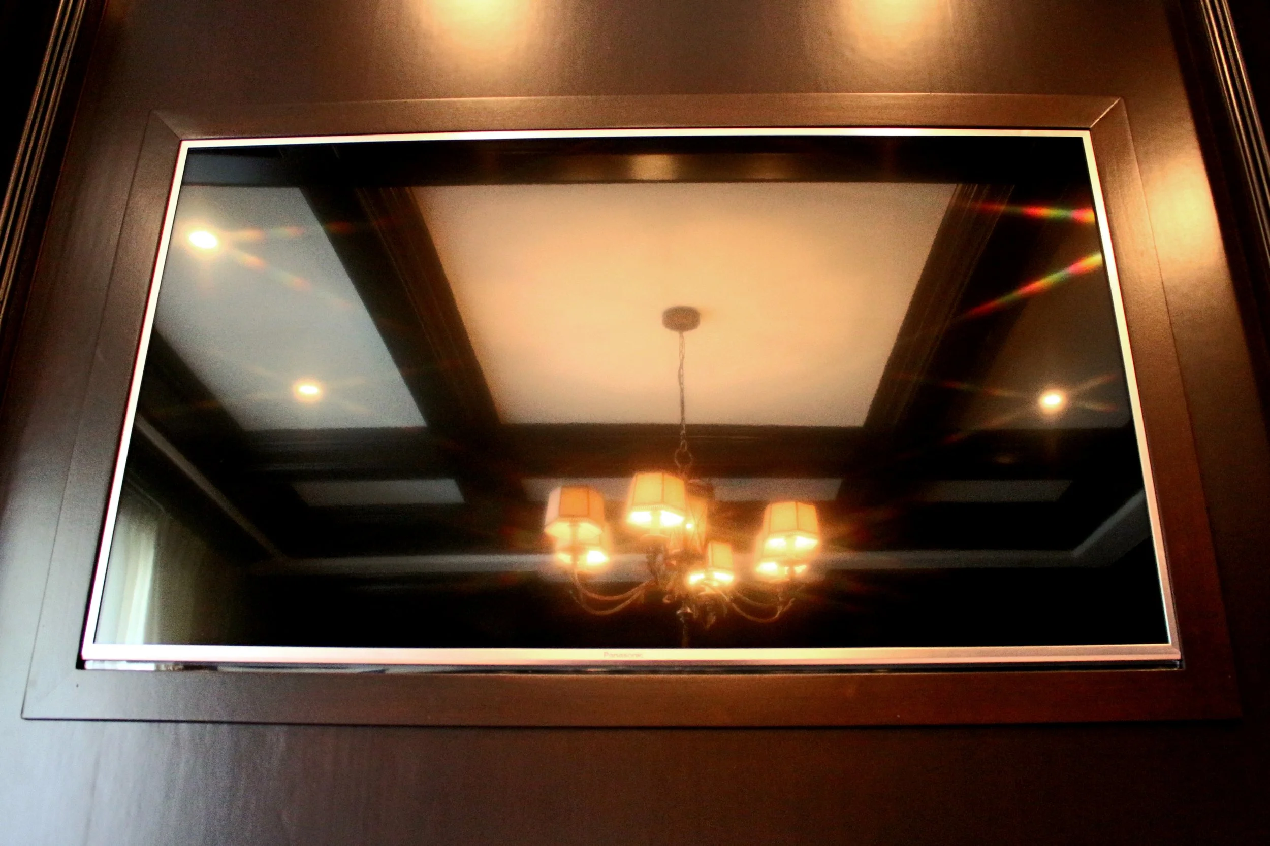 A flat-screen television with a black bezel is turned off, reflecting a ceiling with recessed lighting and a chandelier with multiple lampshades. The TV is placed on a dark wooden surface.