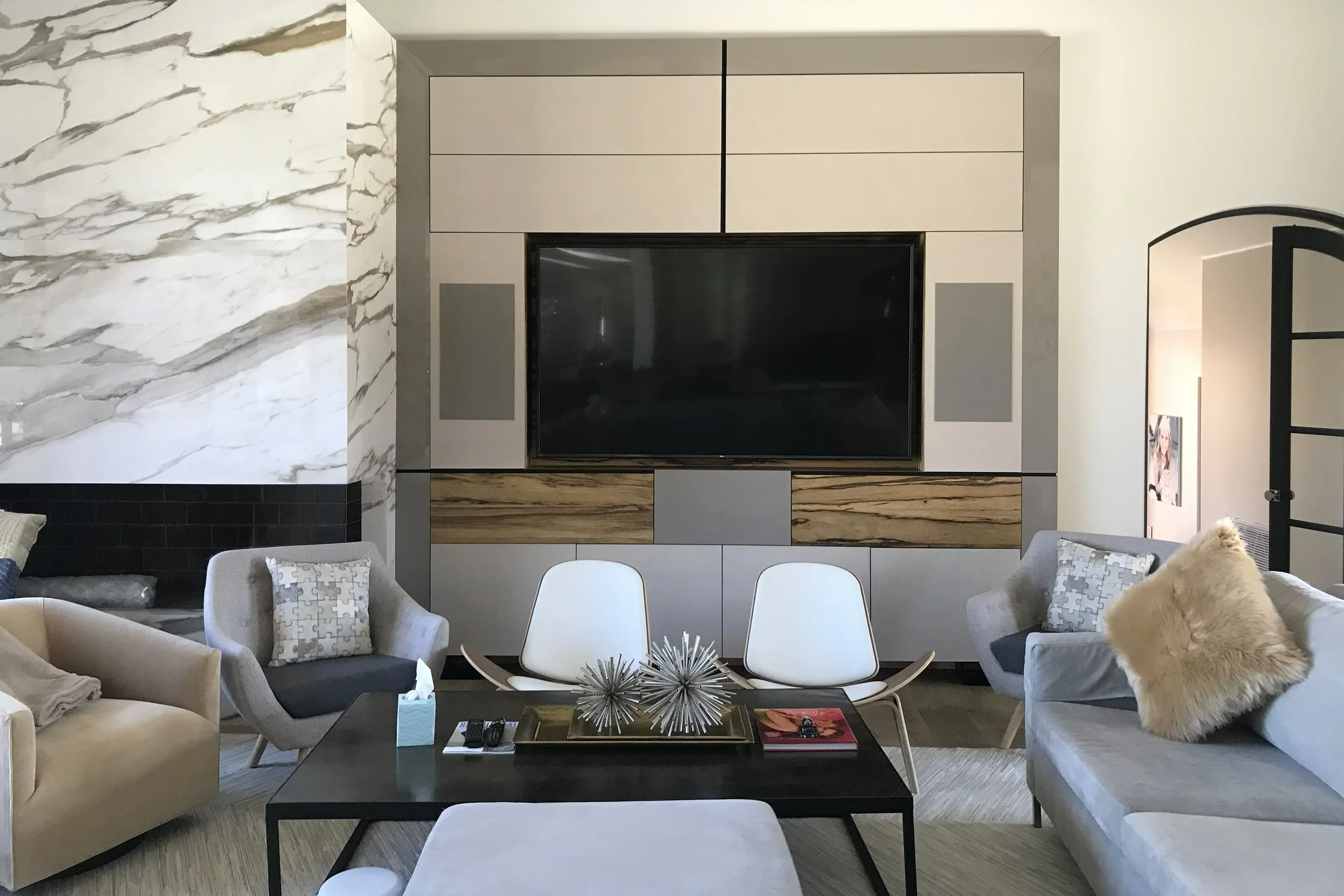 Modern living room with cream and gray furniture, a wall-mounted flat-screen TV, decorative pillows, a black coffee table with books and sculptures, and marble accents on the wall.