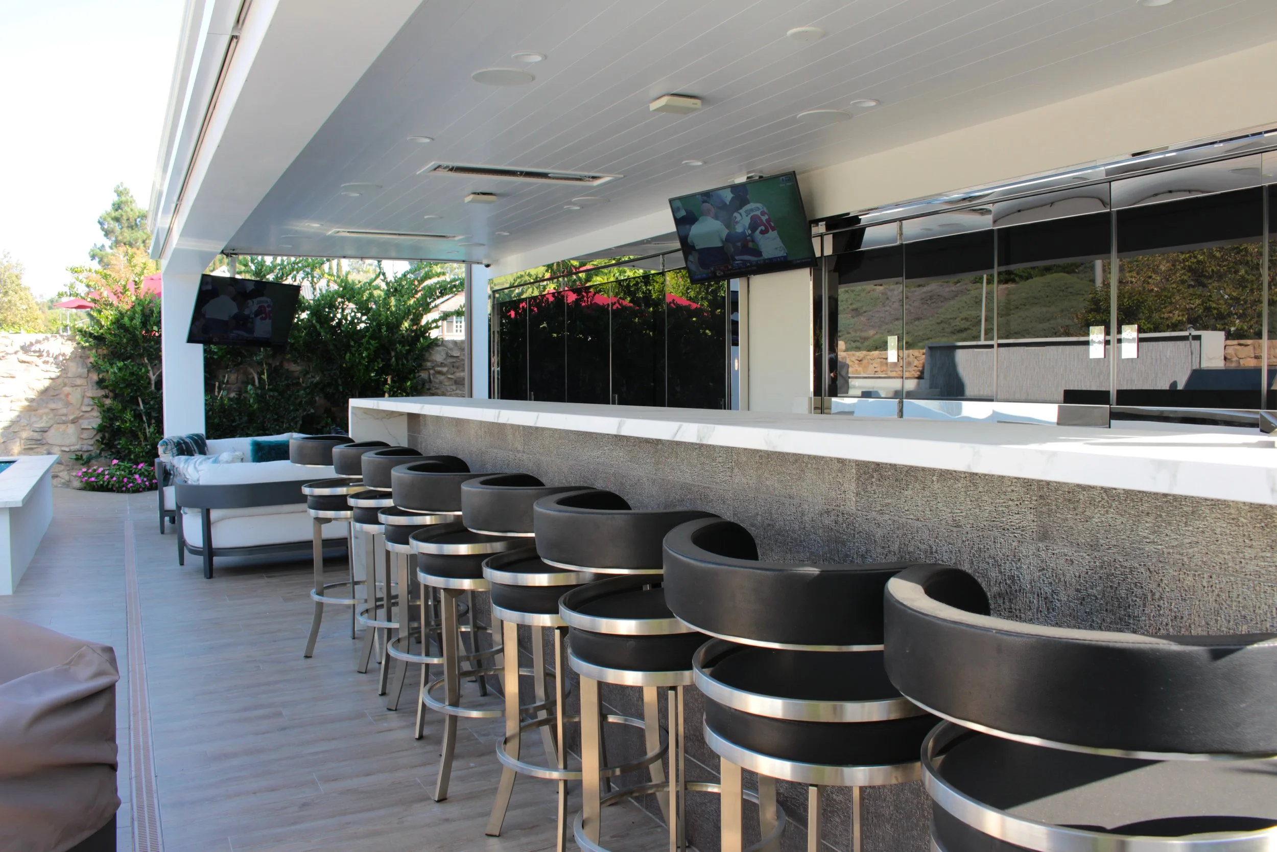 An outdoor bar with black bar stools, a white countertop, and two mounted TV screens, surrounded by greenery and patio furniture.