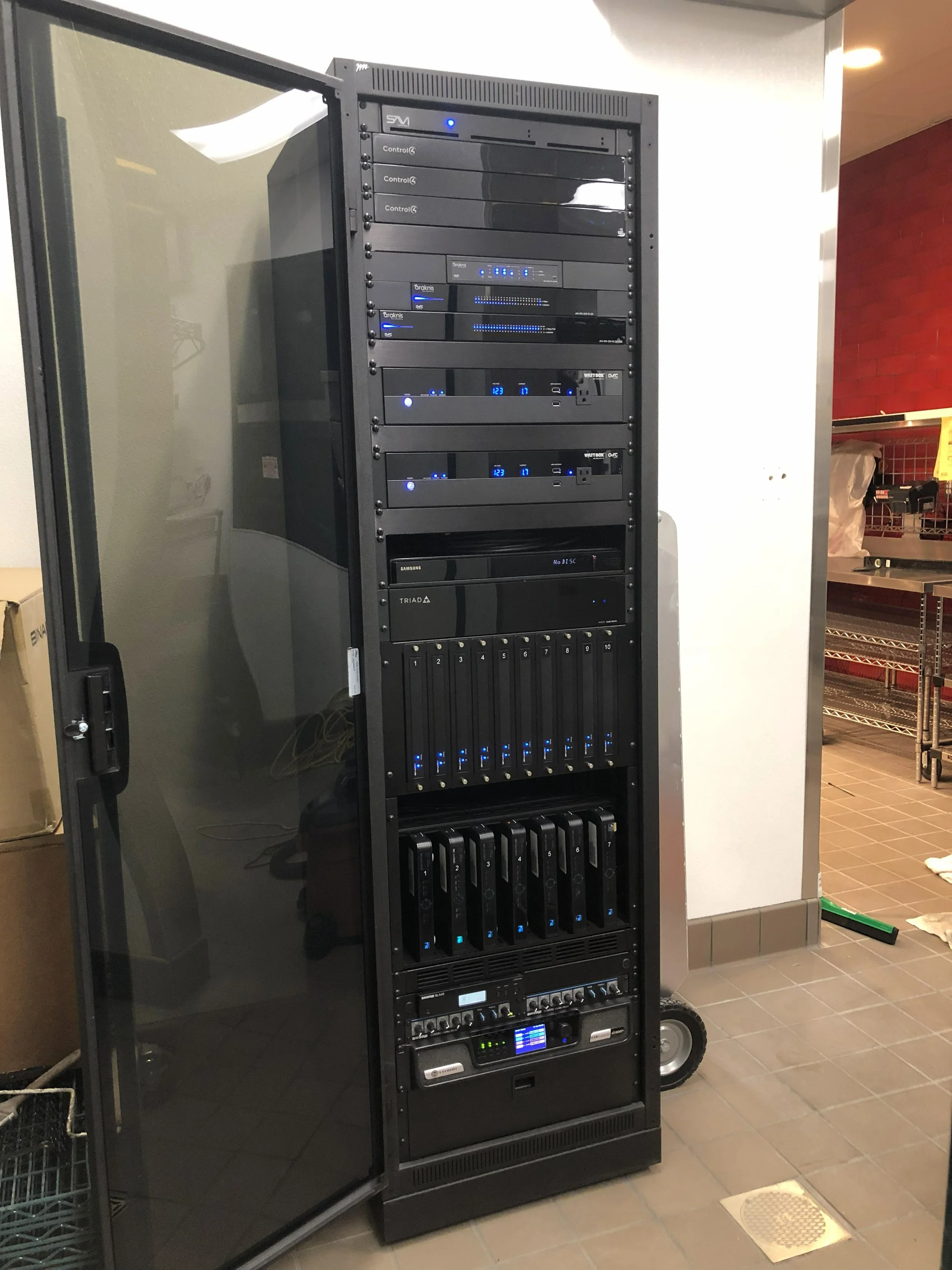 A black server rack containing various electronic equipment, including switches, controllers, and power supplies, with blue LED lights, situated next to a glass door in a tiled room.