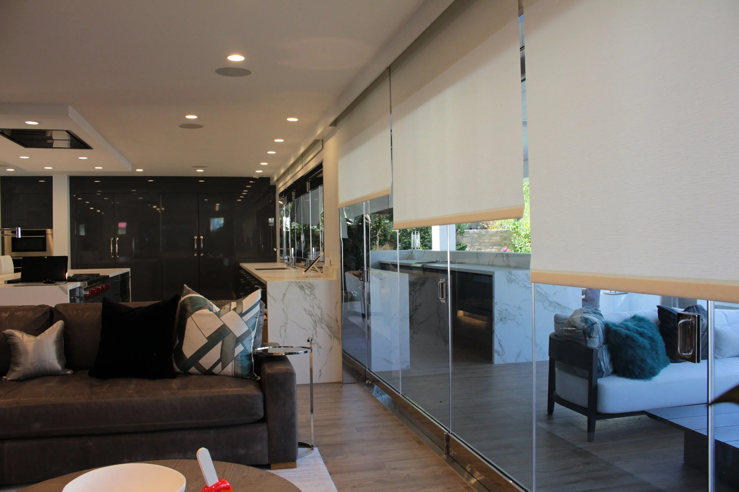 Modern open-concept kitchen and living area with large windows covered by roller shades, black cabinetry, marble countertops, and a brown leather sofa with patterned pillows.