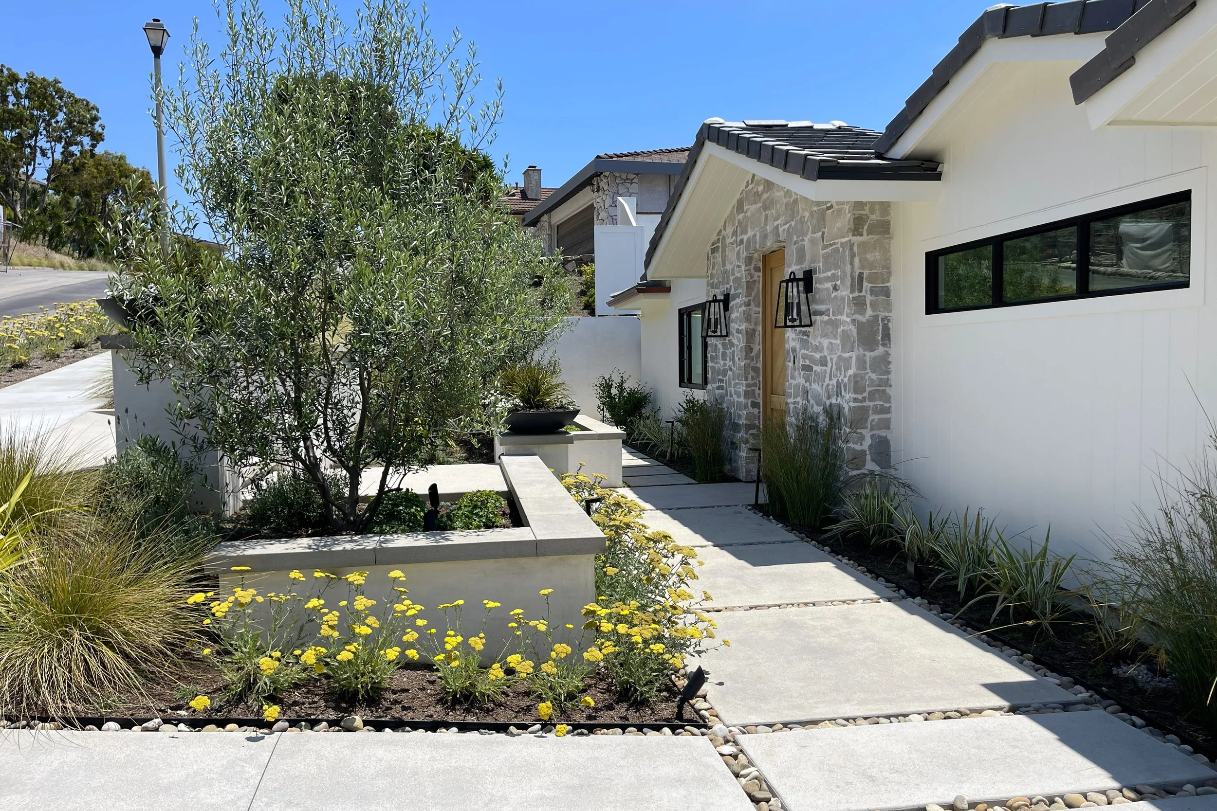 A modern house with stone and white exterior walls, black window frames, and a dark tiled roof, surrounded by landscaped greenery and a clear blue sky.