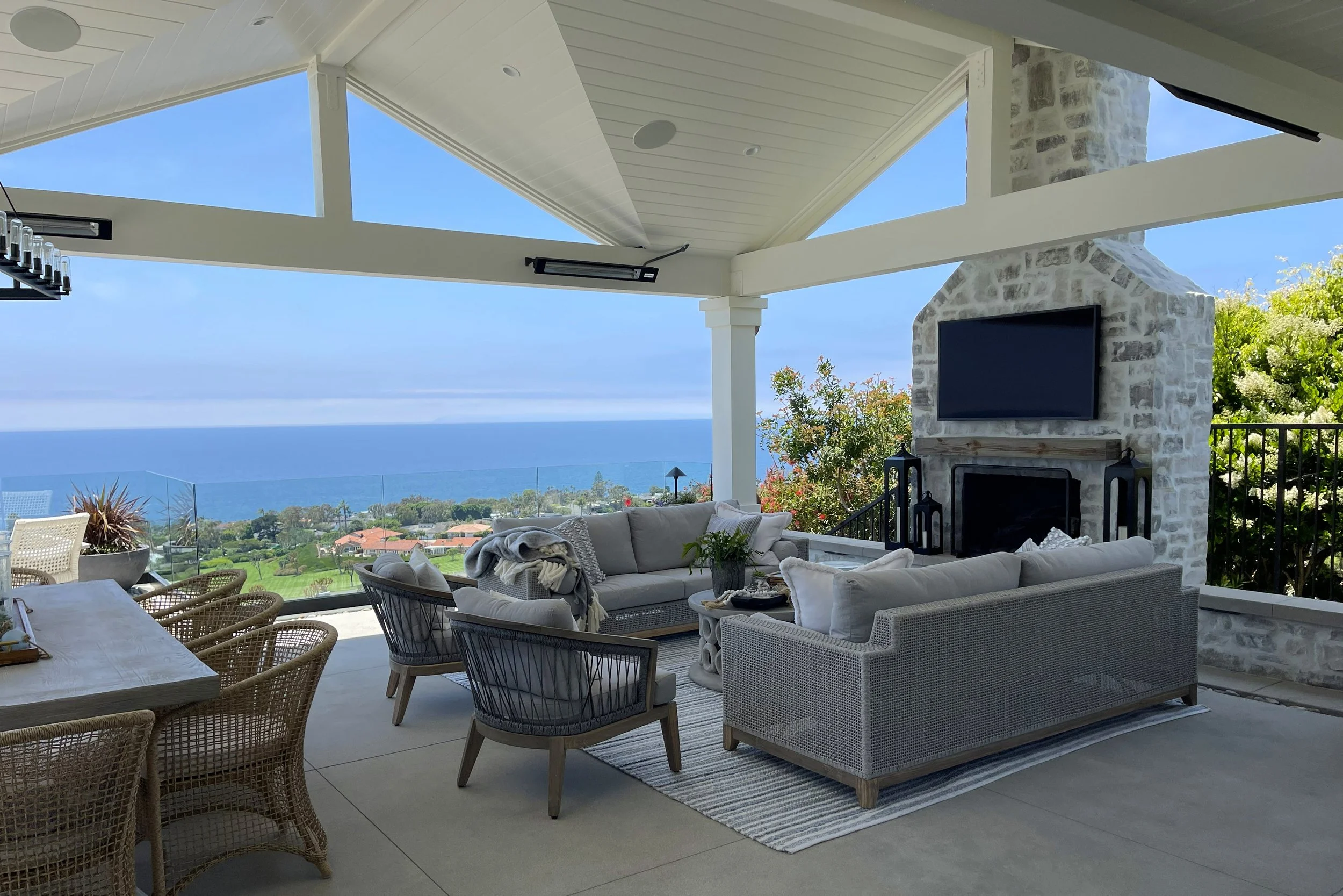 Outdoor patio with white wicker furniture, a stone fireplace with a mounted TV, and ocean view in the background.