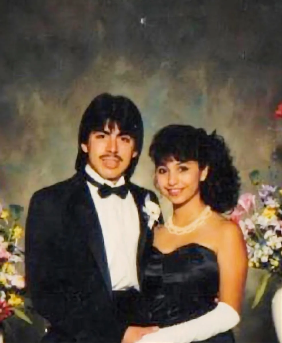 A man and a woman dressed in formal attire, standing together in front of a floral backdrop. The man is wearing a black tuxedo with a bow tie, and the woman is in a strapless black dress with pearl jewelry.