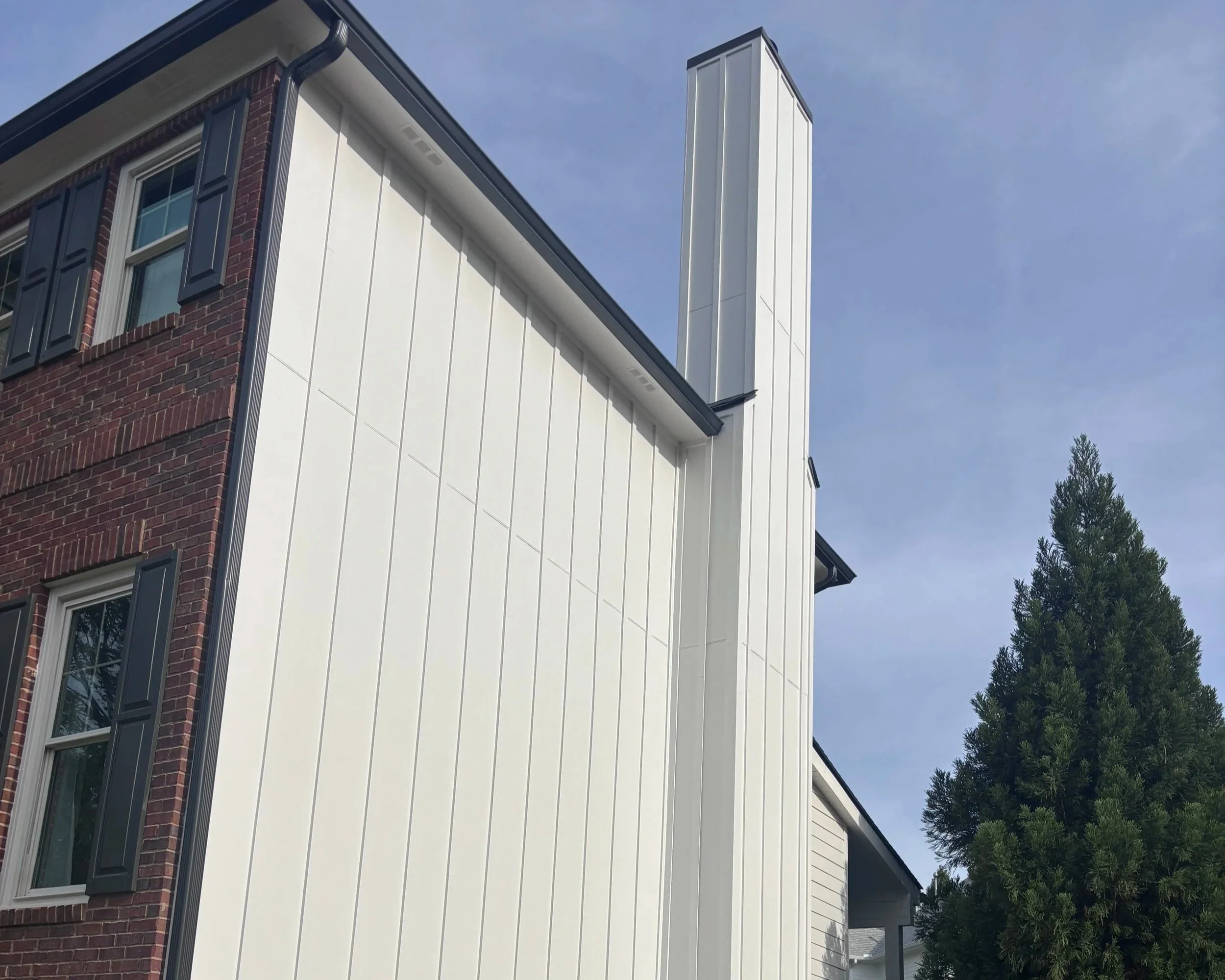 The corner of a multi-story house with a brick facade on one side and white siding on the other. It has several windows with black shutters, a black gutter, and a tall white chimney. The sky is blue with some clouds, and there is a tall evergreen tre