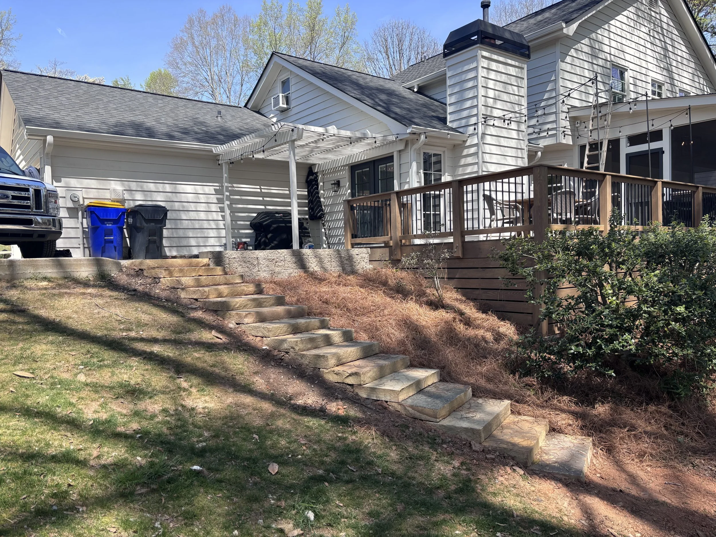 A backyard scene with a white house, a wooden deck, a black BBQ grill, trash bins, stairs leading up to the deck, and a partially covered patio area with string lights.