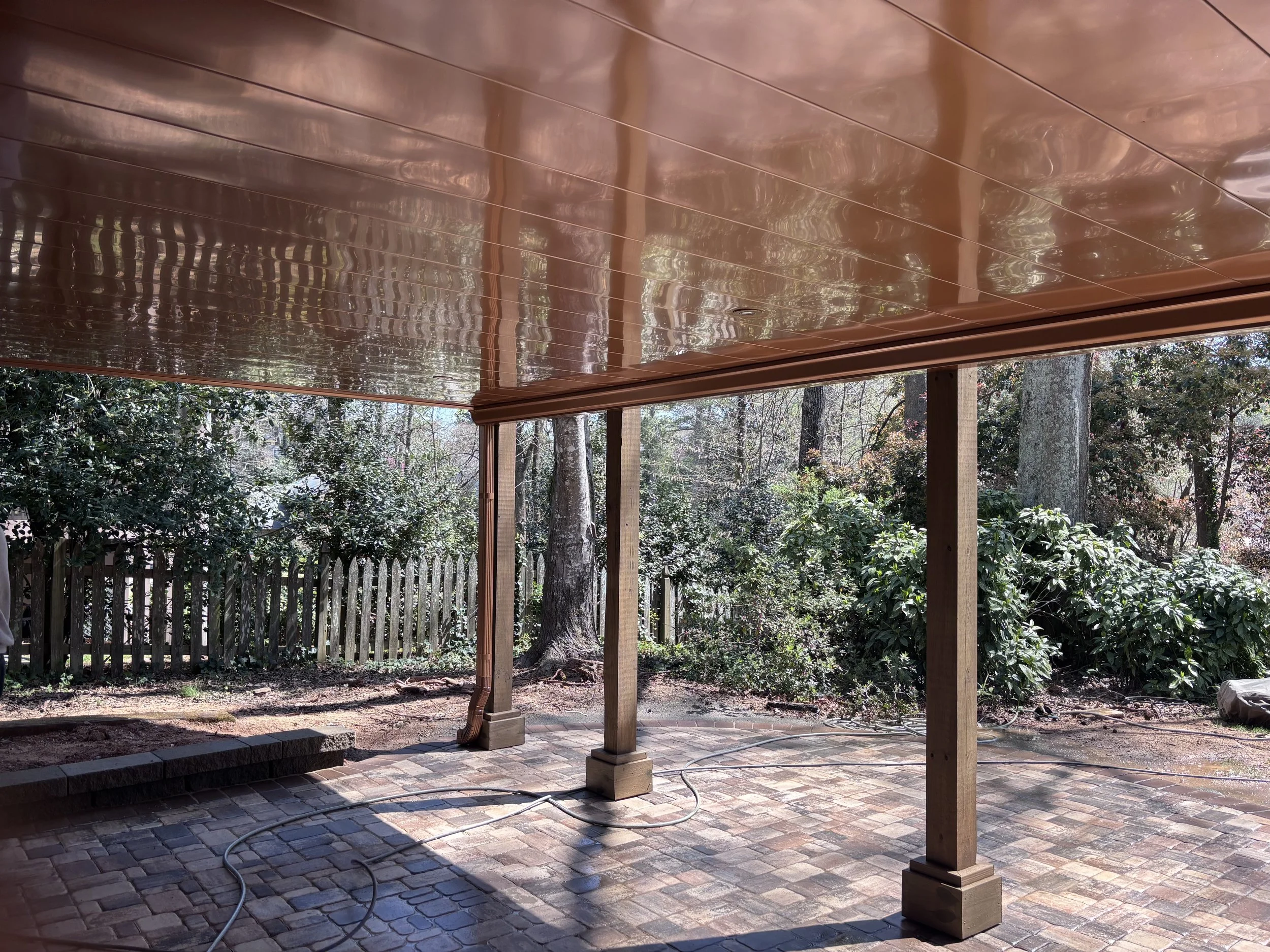 Backyard patio with brick flooring, wooden posts, a new copper-colored roof, surrounded by trees and bushes.