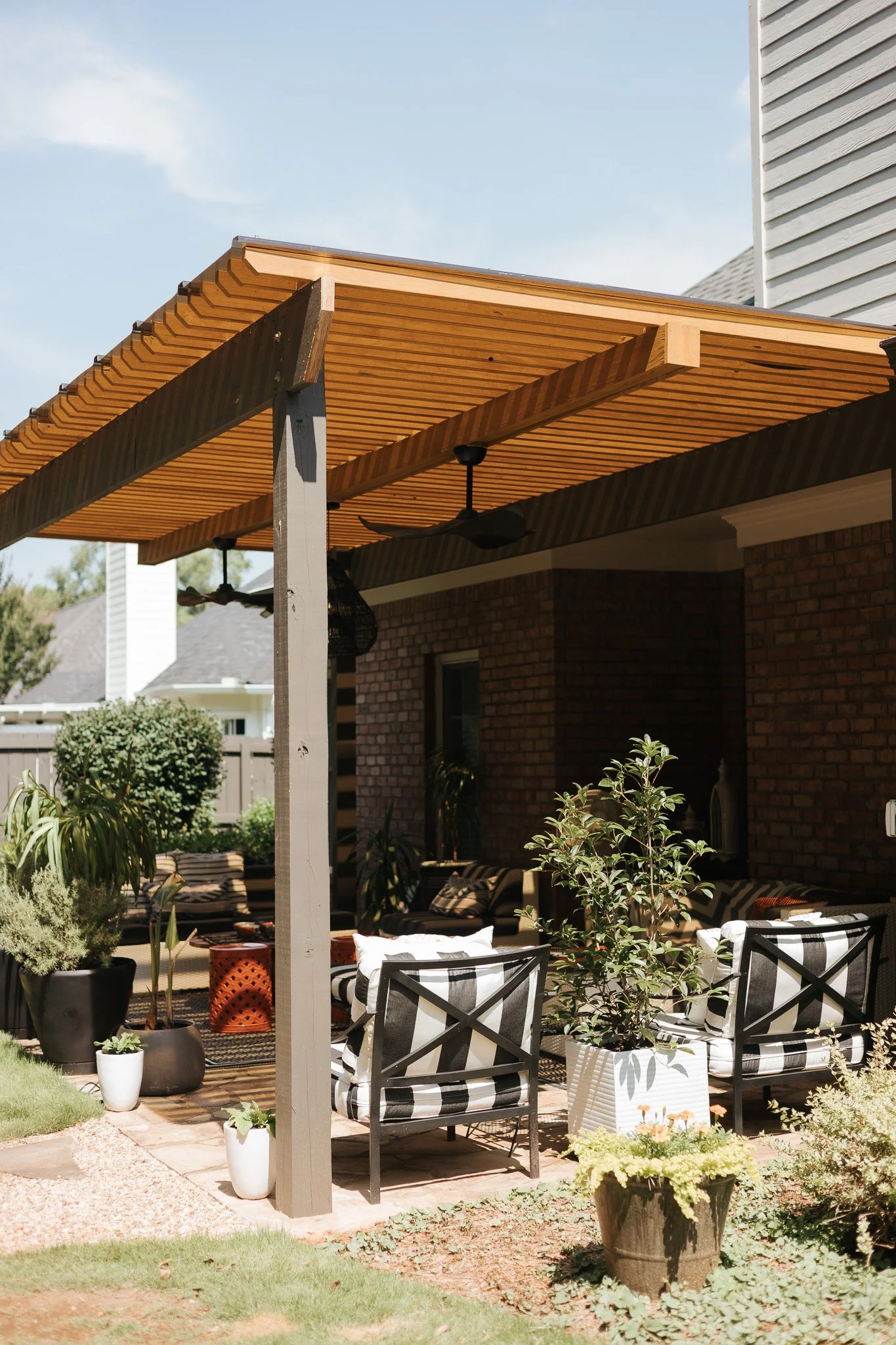 A backyard patio shaded by a wooden pergola with a ceiling fan, surrounded by potted plants, outdoor seating with striped cushions, and a brick house wall.