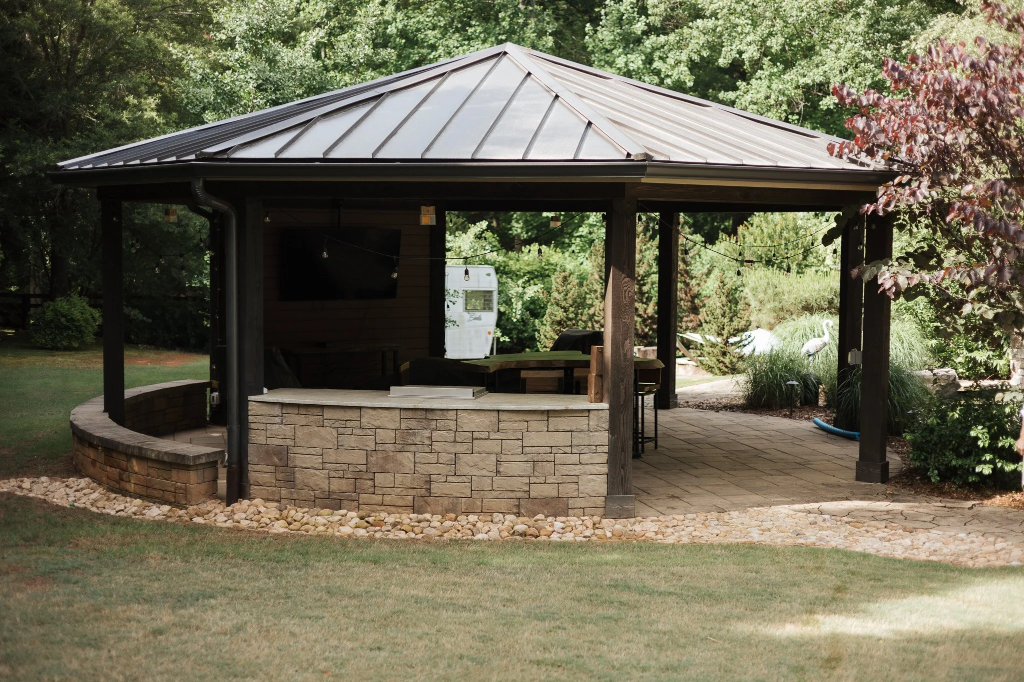 An outdoor living space with a stone fire pit, surrounded by a curved stone border, under a metal-roof gazebo in a garden with trees and greenery.
