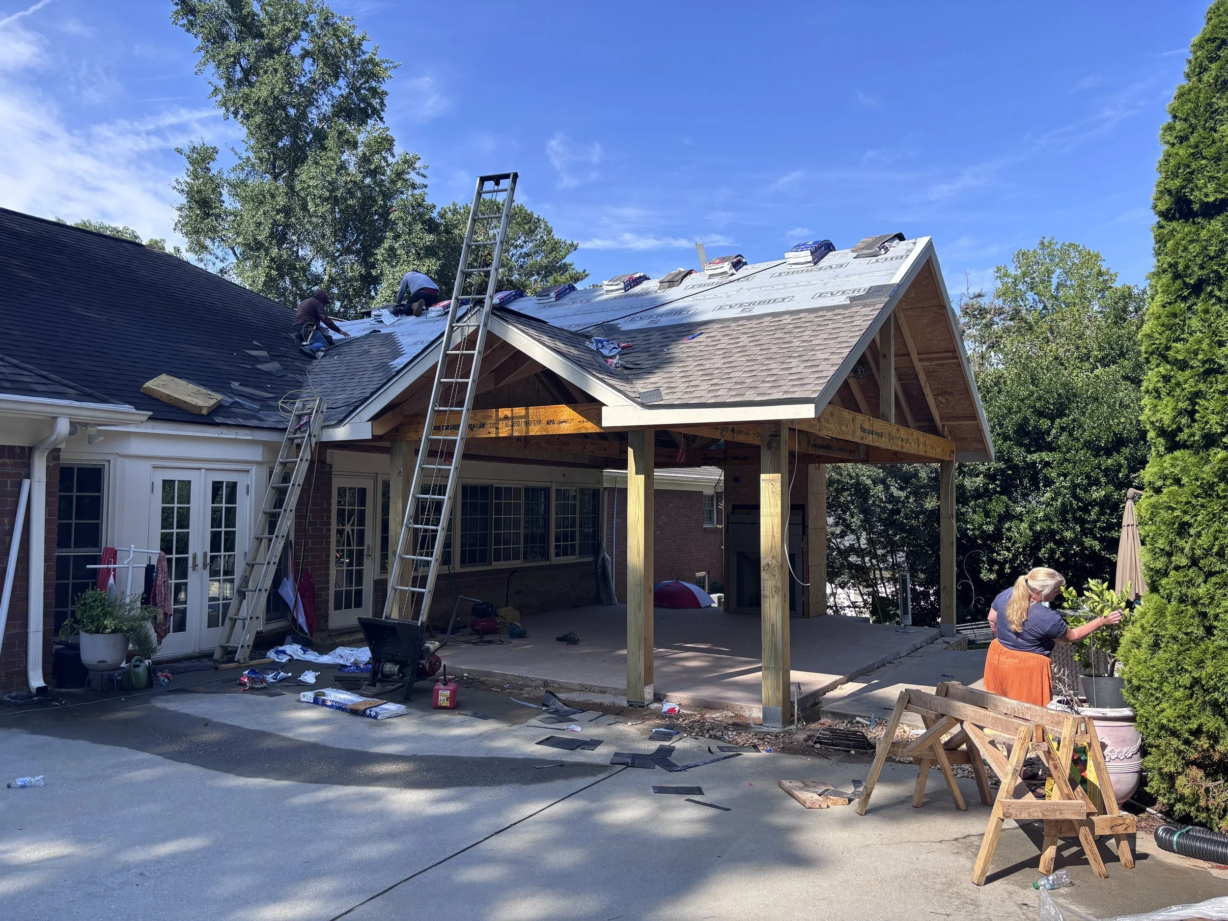 Construction workers installing a roof on a house, with roofing materials spread on the roof and ground, ladders, and a partially built addition with wooden framing under a bright blue sky.