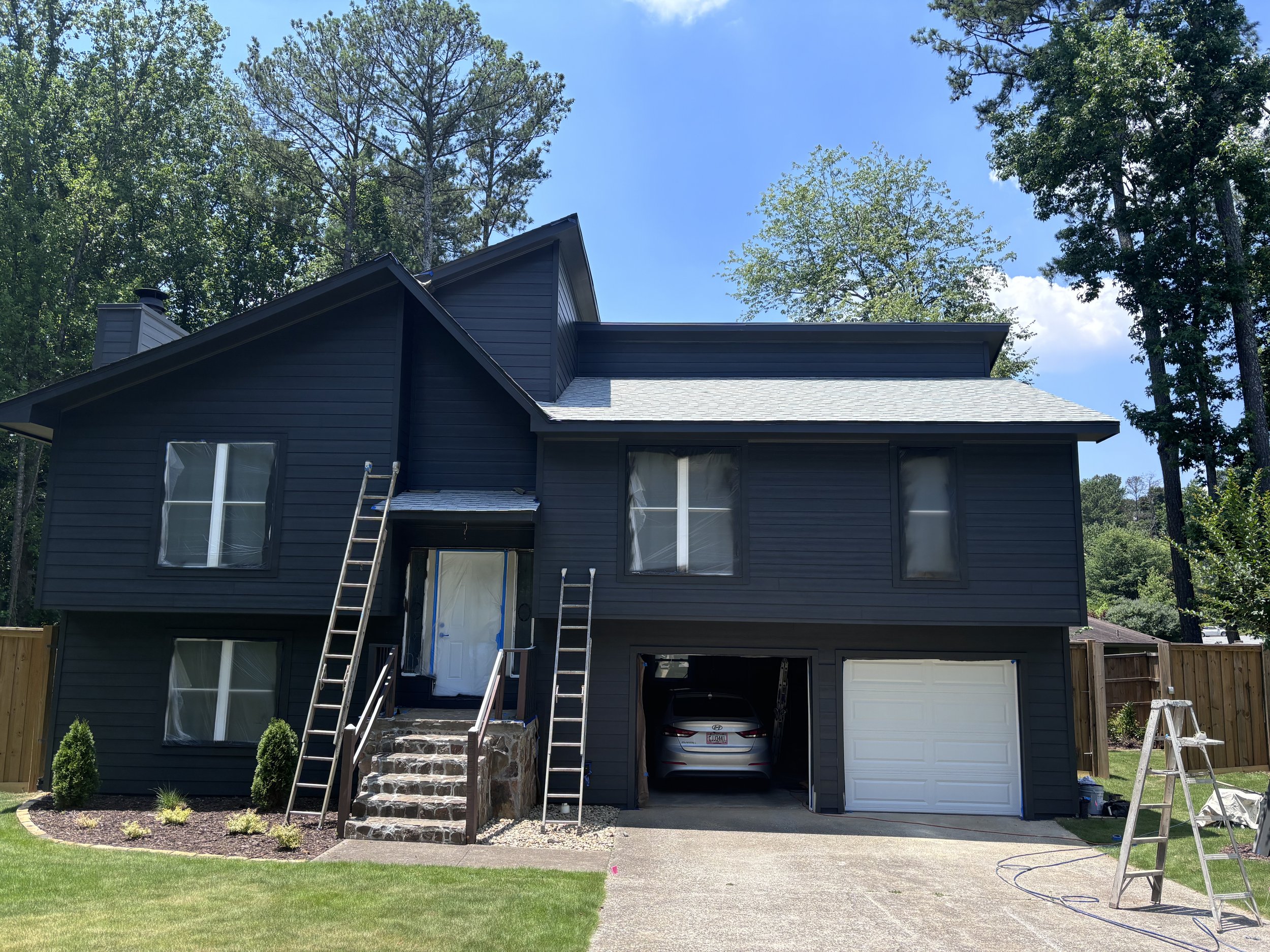 A dark blue two-story house under construction with ladders and tools outside, a partially installed garage door, and surrounded by a grassy yard with small bushes, trees, and a clear blue sky.