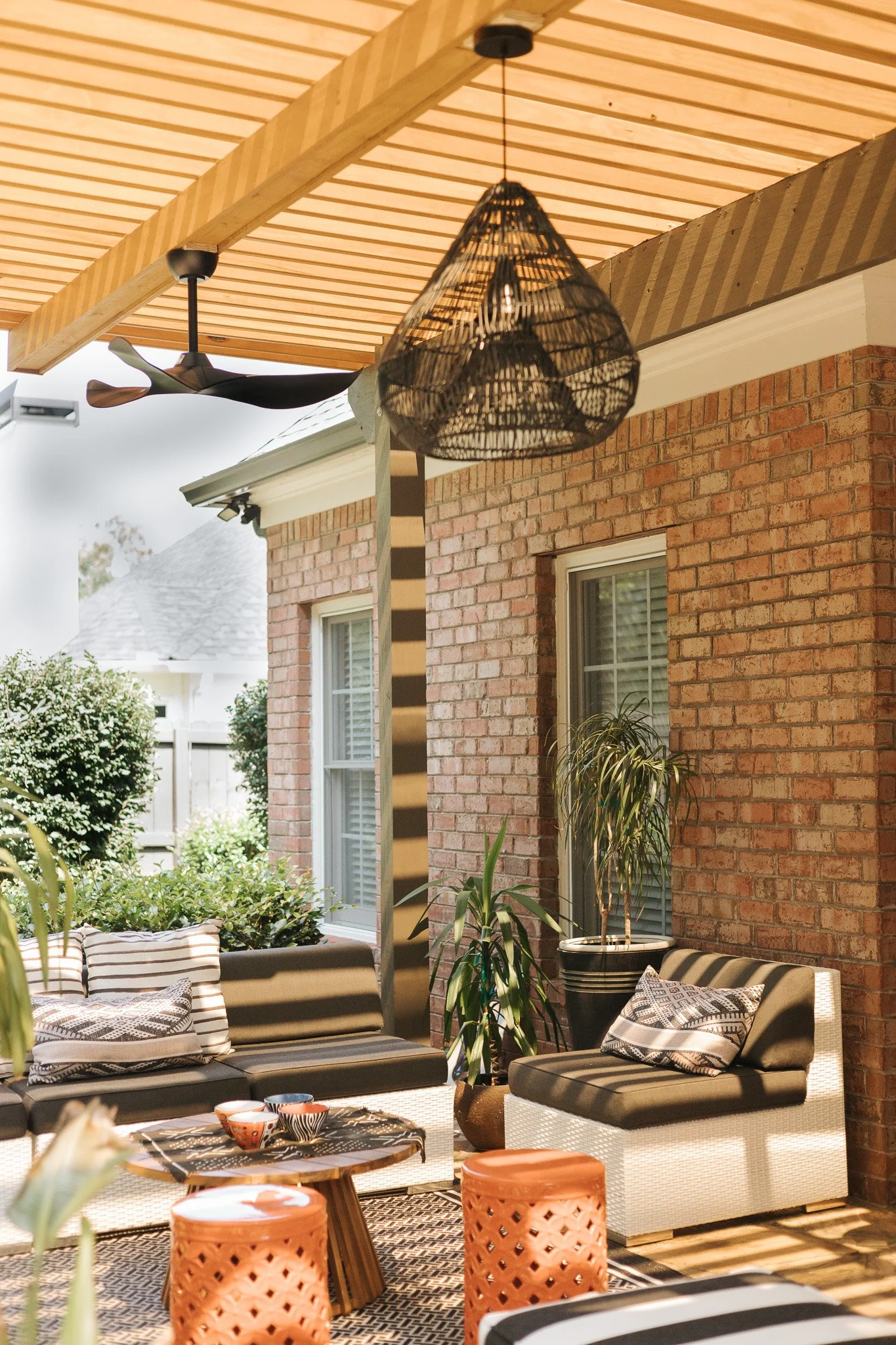 Outdoor patio area with black cushioned seating, striped pillows, potted plants, a hanging wicker light, and a ceiling fan, adjacent to a brick house with large windows.