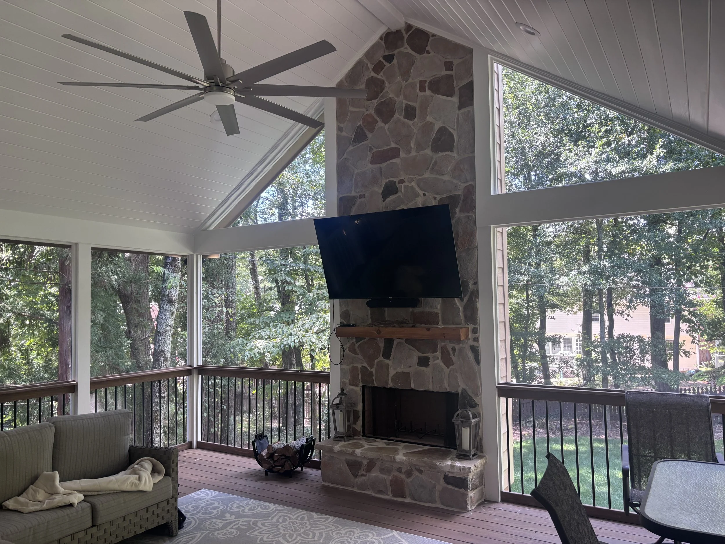 A living room with large windows, a ceiling fan, a stone fireplace, a couch with blankets, and outdoor greenery visible through the windows.